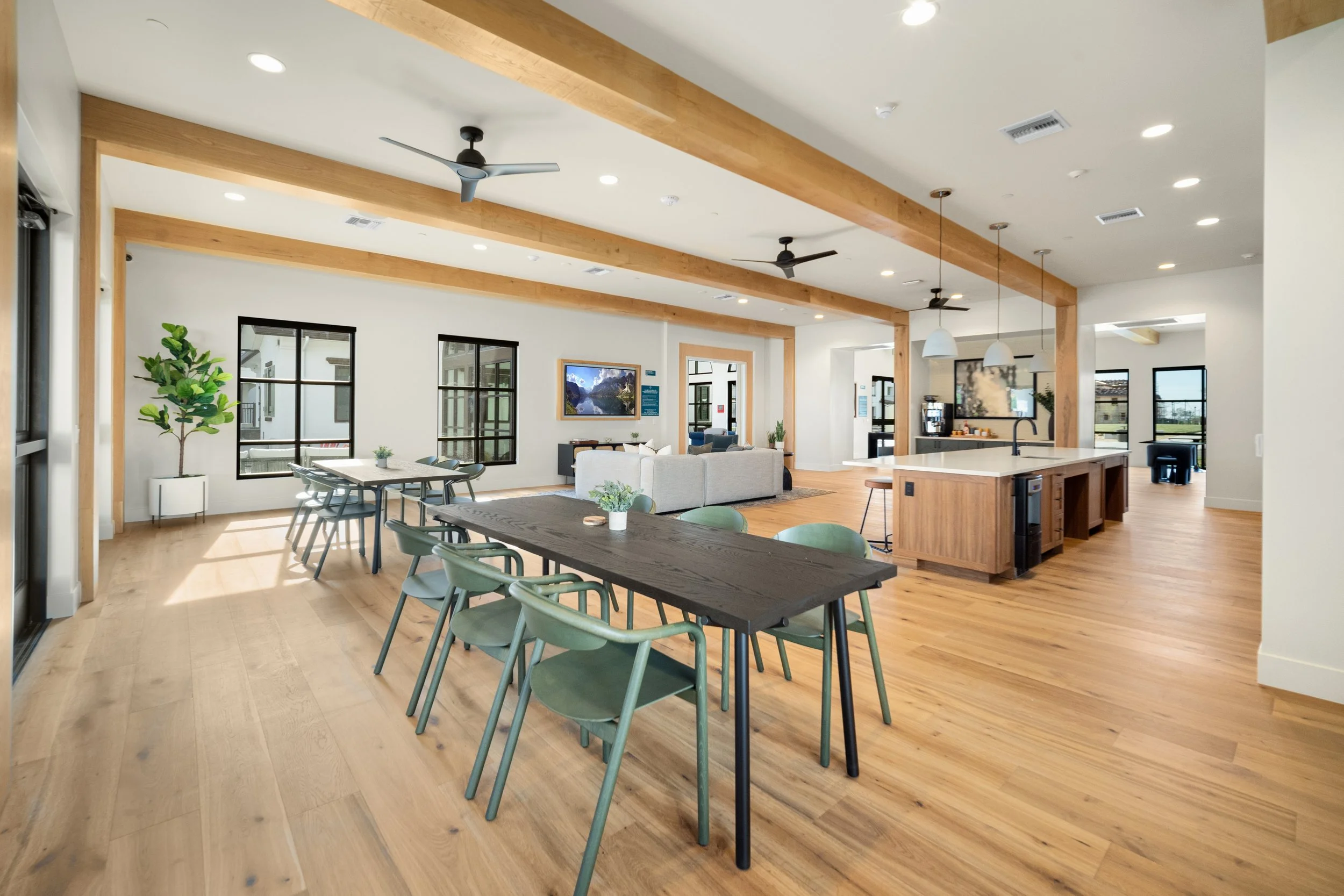 Spacious, modern living room with open floor plan, wooden beams, ceiling fans, large windows, and hardwood flooring. Furnished with dining table, chairs, sofa, and kitchen island.