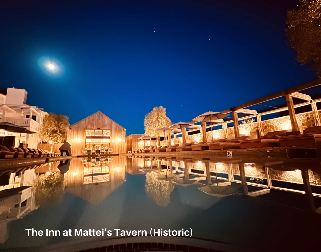 Nighttime view of an outdoor pool area at The Inn at Mattei's Tavern, with wooden buildings, lounge chairs with umbrellas, and a clear sky with the moon and stars reflected in the pool.