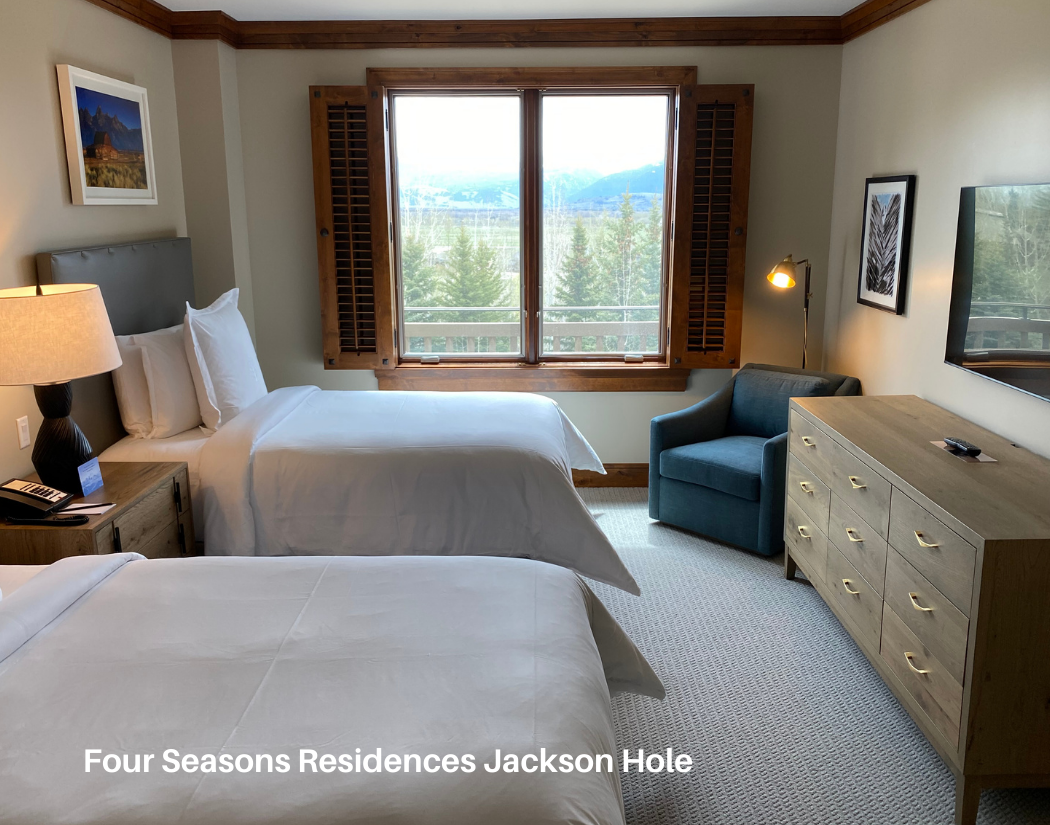 A hotel room with two beds, a wooden dresser, a blue armchair, a window showing trees and mountains, wall art, and a flat-screen TV.