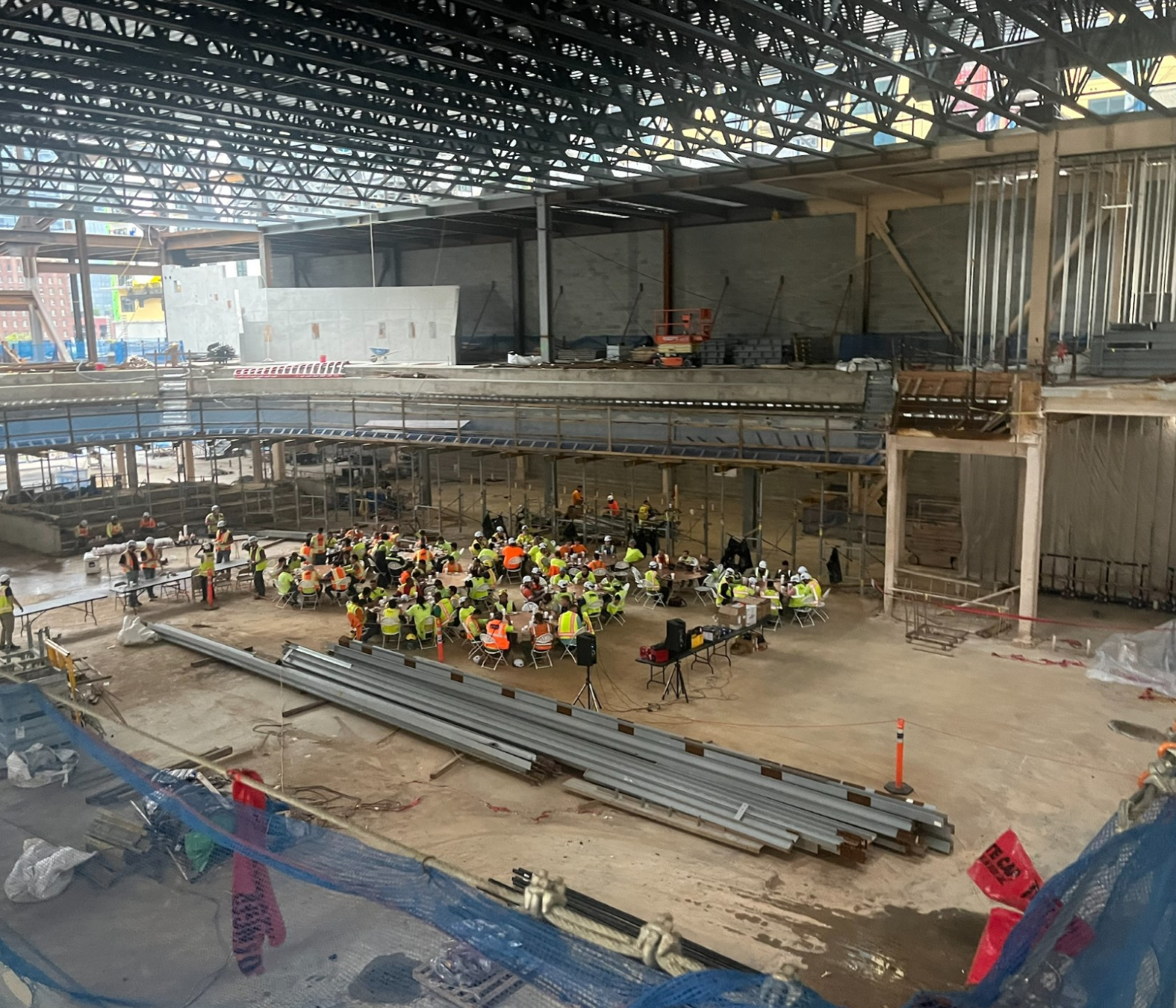 Construction site with workers and equipment, and a group of people sitting at tables inside a large unfinished building.