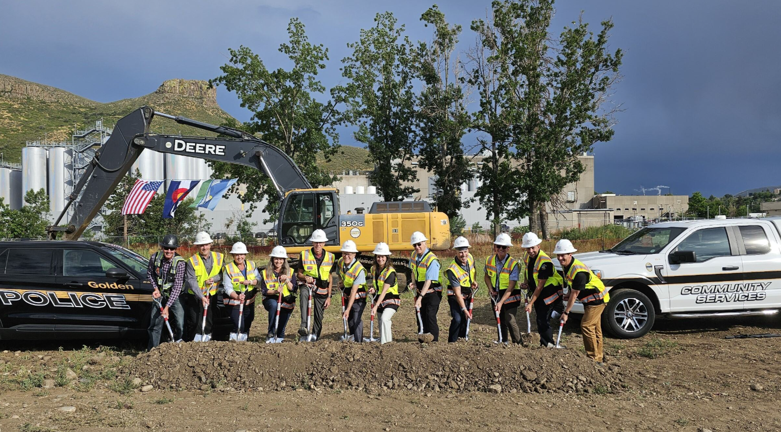 People wearing safety vests and hard hats participating in a groundbreaking ceremony, lining up with shovels in front of police and community services vehicles, with construction equipment and trees in the background.