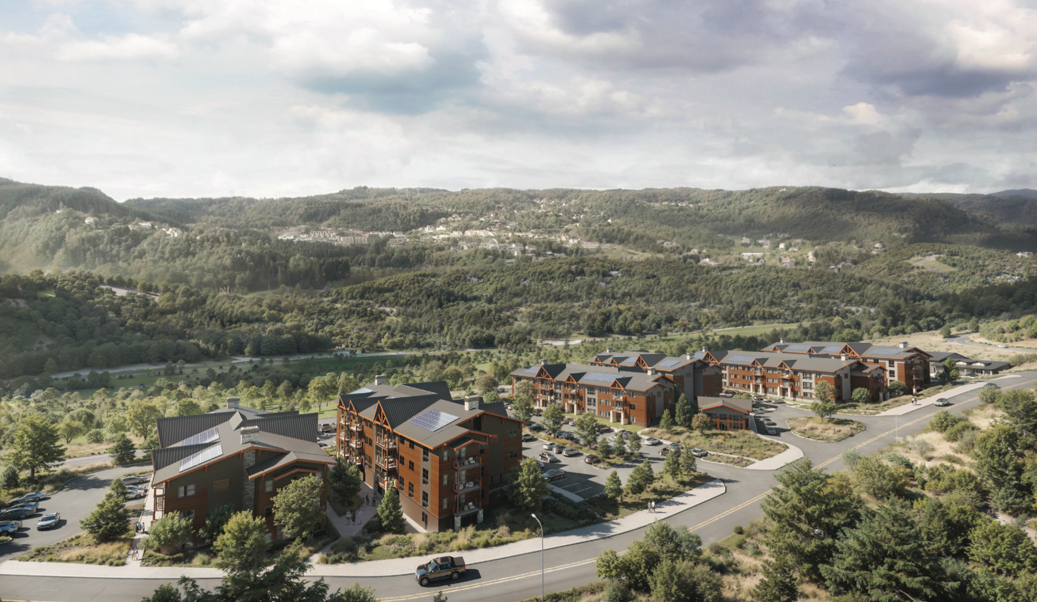 Aerial view of a residential complex with multiple buildings, parking lots, surrounded by trees, hills, and mountains in the background, under a partly cloudy sky.