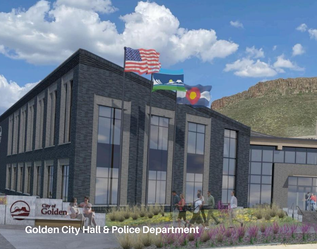 A modern government building with three flags flying on top: the United States flag, the state flag of Washington, and the flags of the City of Golden. People are walking and sitting outside, and there are landscaped plants around the building. A sig