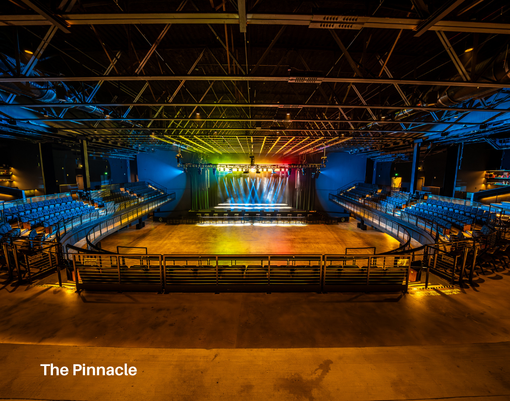 An empty indoor concert or event venue with a stage equipped with colorful stage lighting and an empty floor in front. Seating arrangements are on both sides of the stage with blue chairs, and the venue has high ceilings with visible metal structural