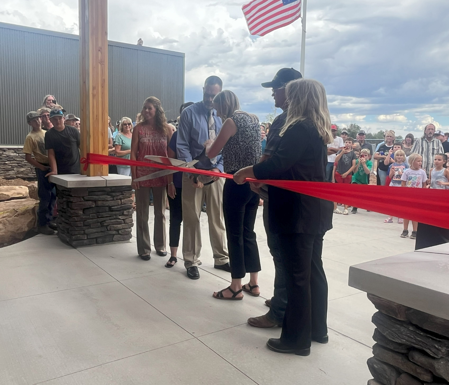 A ribbon-cutting ceremony outdoors with a crowd of people, officials, and children, with an American flag and cloudy sky in the background.