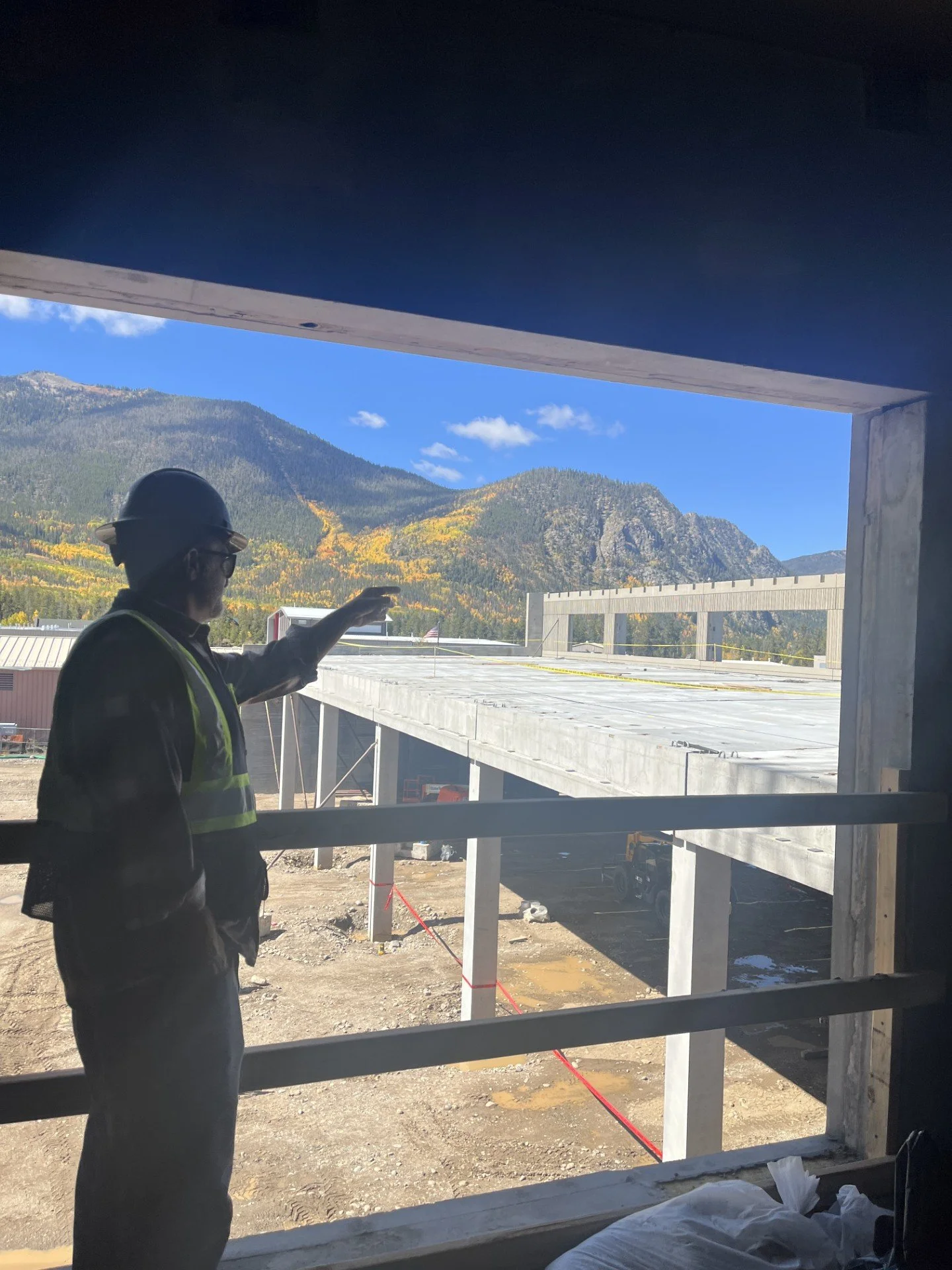 Construction worker wearing a safety helmet and vest looking out of a window at a bridge under construction with mountainous landscape and blue sky in the background.