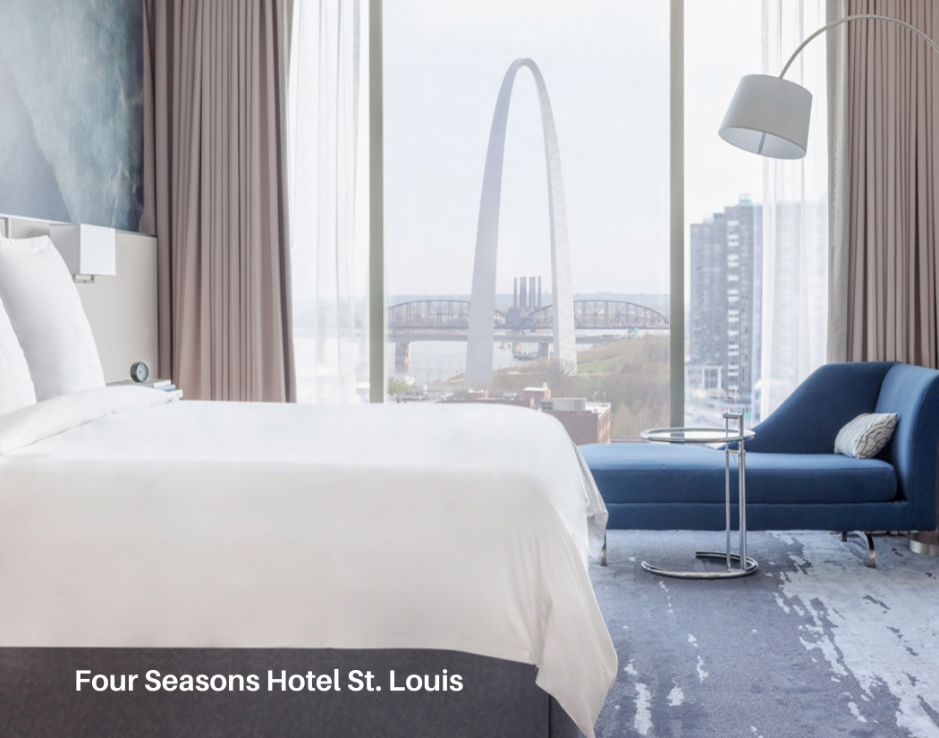 Hotel room with a large window view of the St. Louis Gateway Arch and Mississippi River, featuring a white bed, blue chaise lounge, floor lamp, and beige curtains.