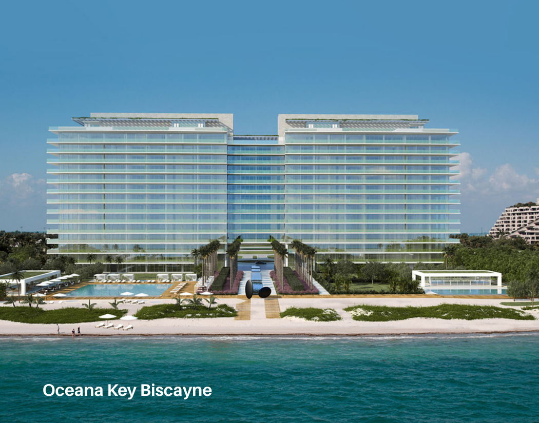 A large modern beachfront hotel, Oceana Key Biscayne, with multiple floors, glass facade, an outdoor swimming pool, lounge chairs, palm trees, and a sandy beach in front. The ocean is visible in the foreground under a clear blue sky.