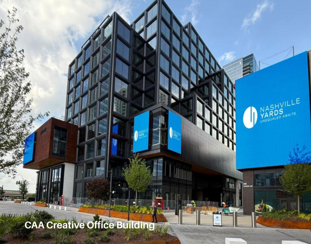 Exterior view of the CAA Creative Office Building with modern glass and steel architecture, large digital screens displaying Nashville Yards advertisements, and landscaped surroundings, under a partly cloudy sky.