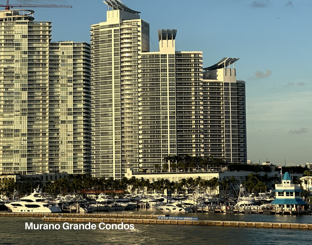 High-rise condos overlooking a marina with docked boats, palm trees, and a blue sky with some clouds, labeled as Murano Grande Condos in Miami Beach.