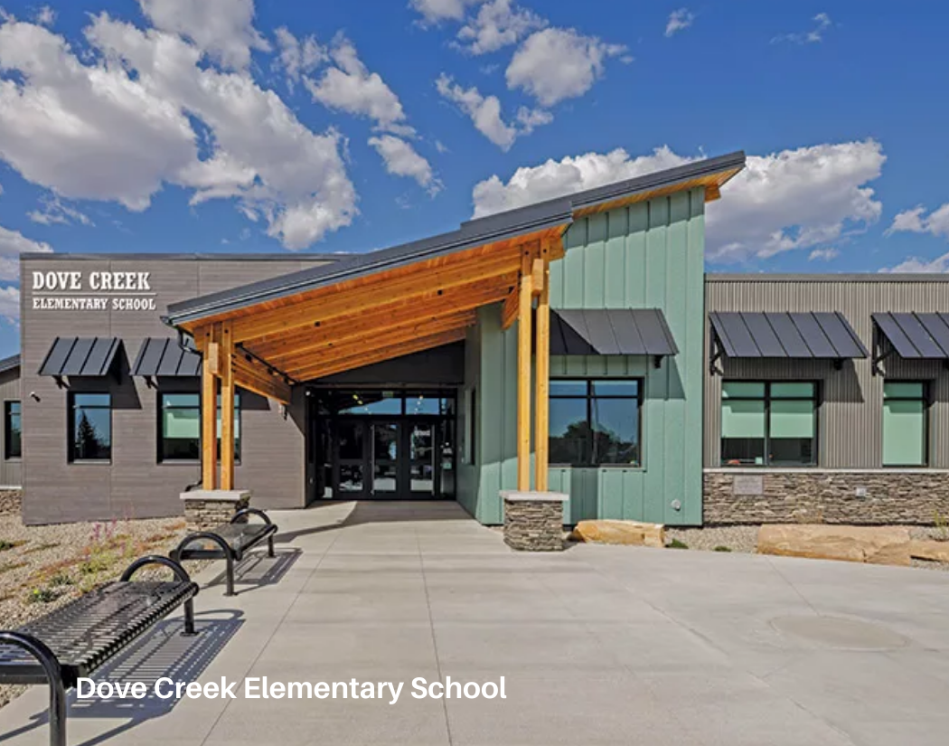 The modern exterior of Dove Creek Elementary School with a welcoming entrance, benches, and a partly cloudy sky.