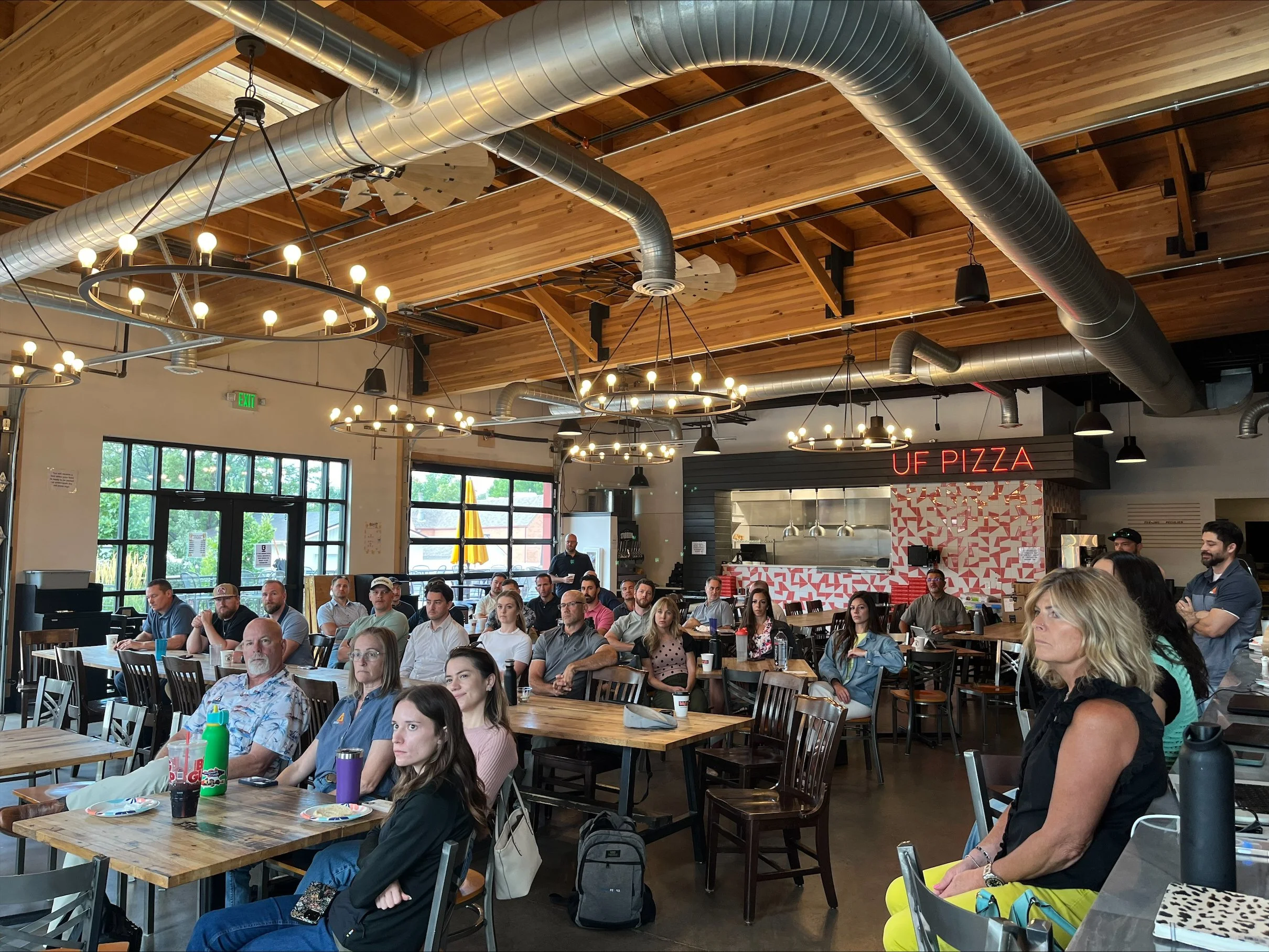 People seated at tables inside a restaurant with a modern industrial design, including exposed ductwork, wooden ceiling, and large windows. A sign reads 'UF PIZZA' in neon above the open kitchen area.
