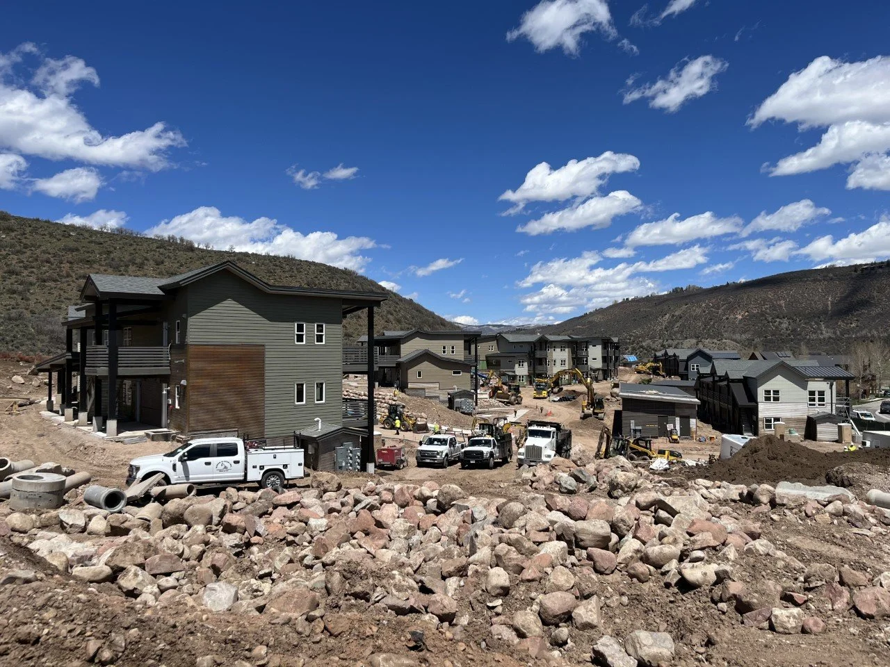 Construction site with new houses being built, construction vehicles, and rocky terrain under a blue sky with clouds.