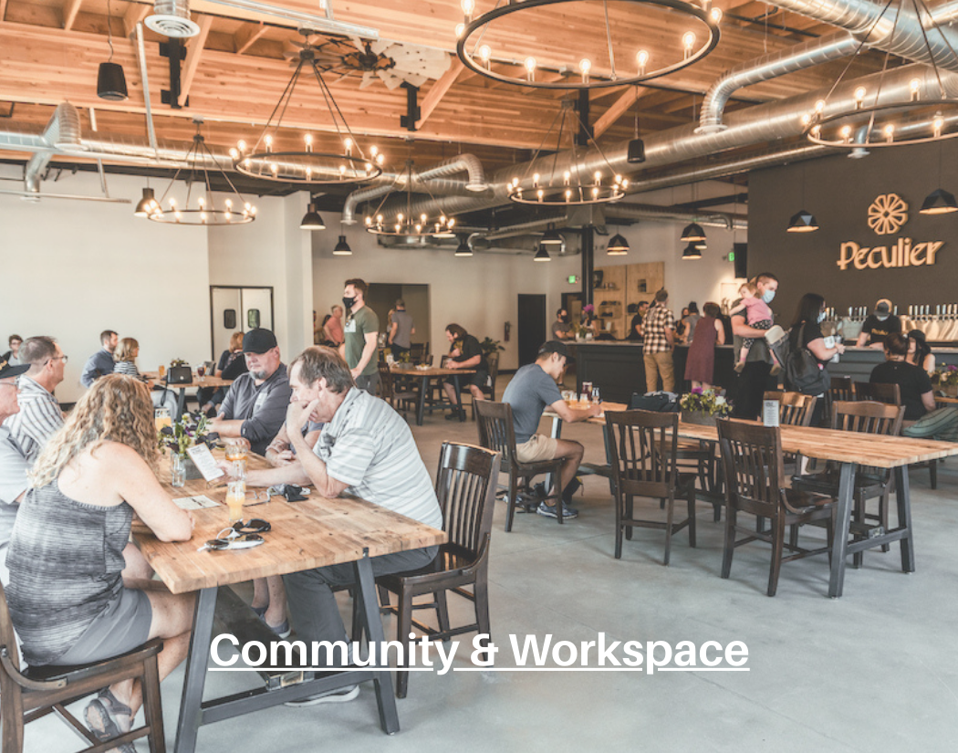 People sitting and working at wooden tables inside a modern, industrial-style community and workspace with high ceilings, exposed ductwork, and hanging light fixtures, a counter area in the background with people ordering drinks, and a sign that says