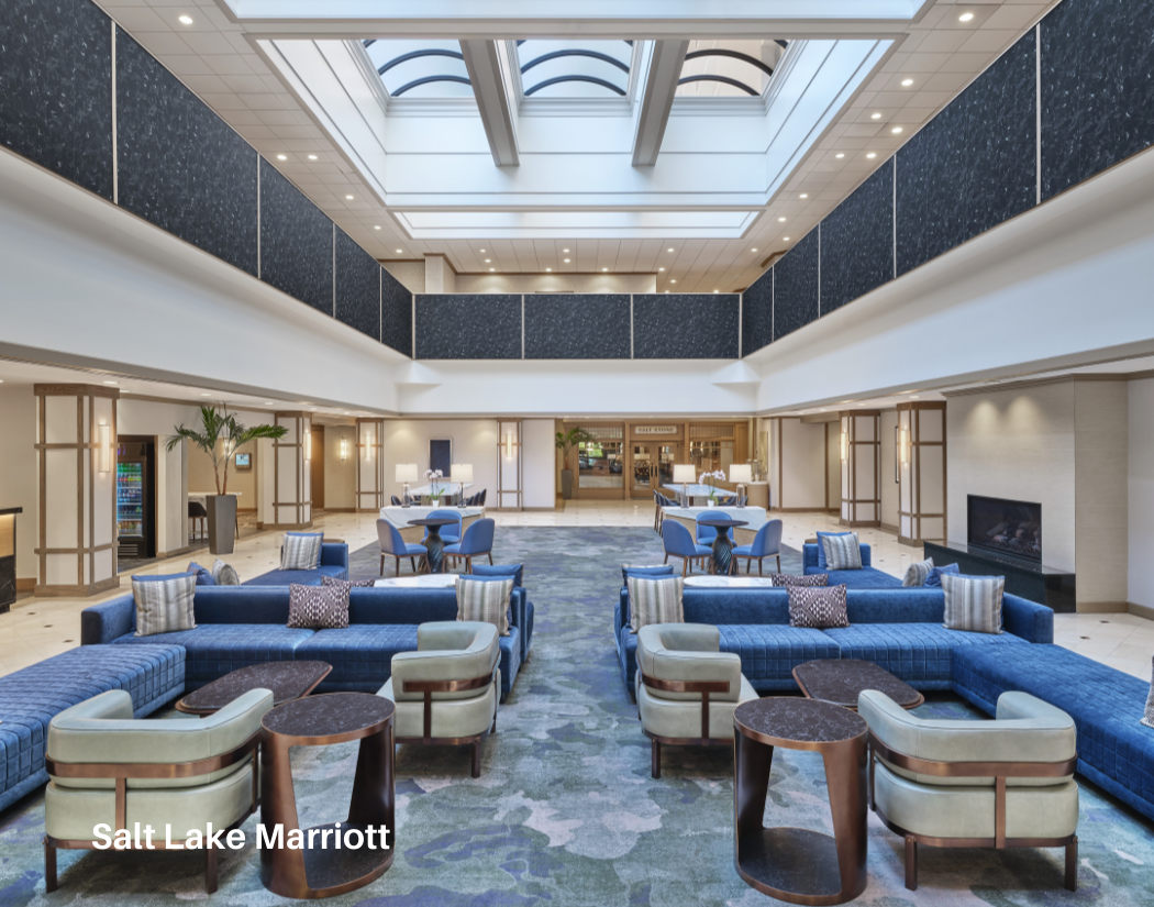 Spacious hotel lobby with blue couches, beige armchairs, circular coffee tables, and a patterned carpet. Tall plants, lamps, and a fireplace are visible, with large skylights and a second-floor balcony overlooking the area. Salt Lake Marriott