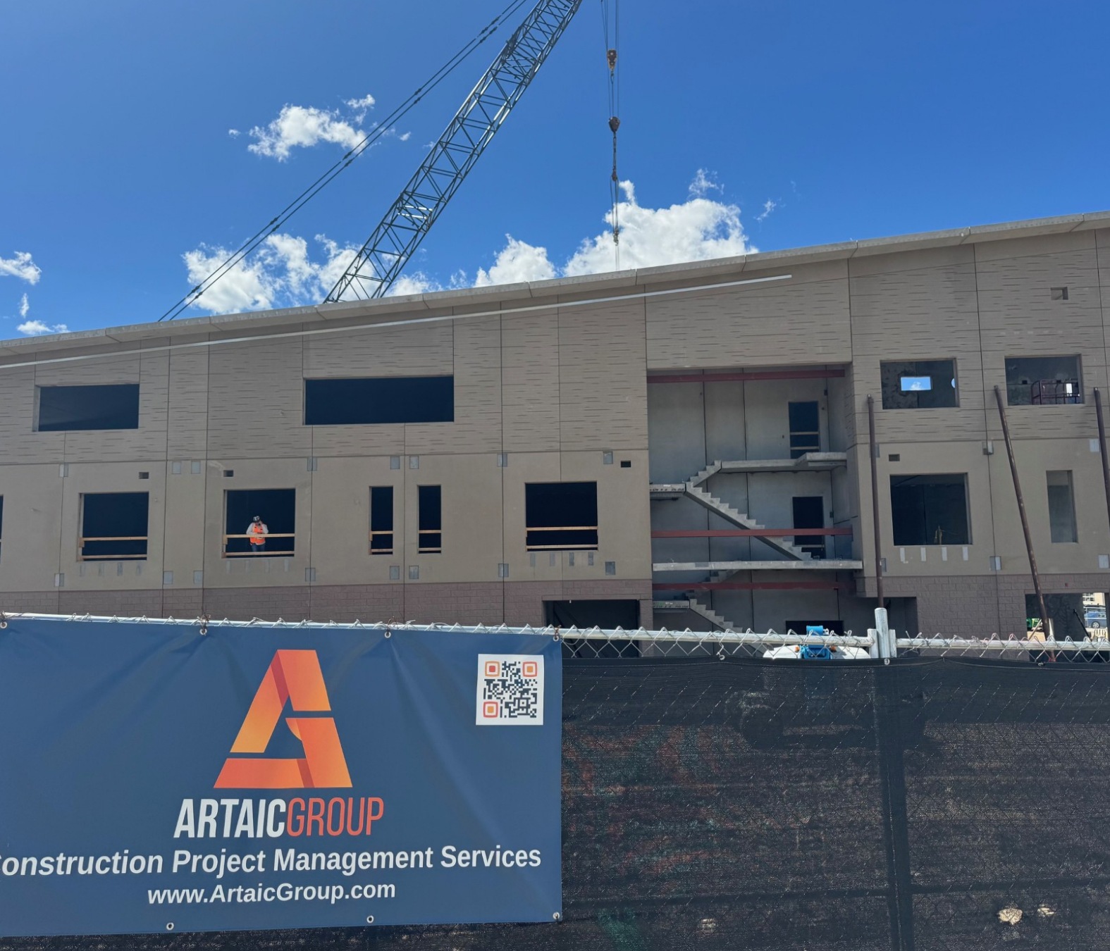 Construction site with a building under construction, a crane overhead, and a fence with a banner displaying the Artaic Group logo and a QR code.