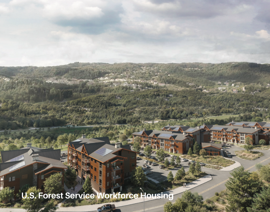 A residential housing complex with multi-story buildings and parking lots, surrounded by lush greenery and mountains in the background, under cloudy skies.