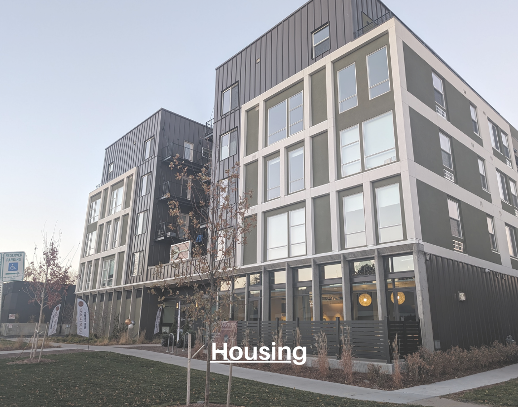 Modern multi-story apartment building with large windows and outdoor balconies, surrounded by a landscaped area with young trees and a reserved handicap parking spot.
