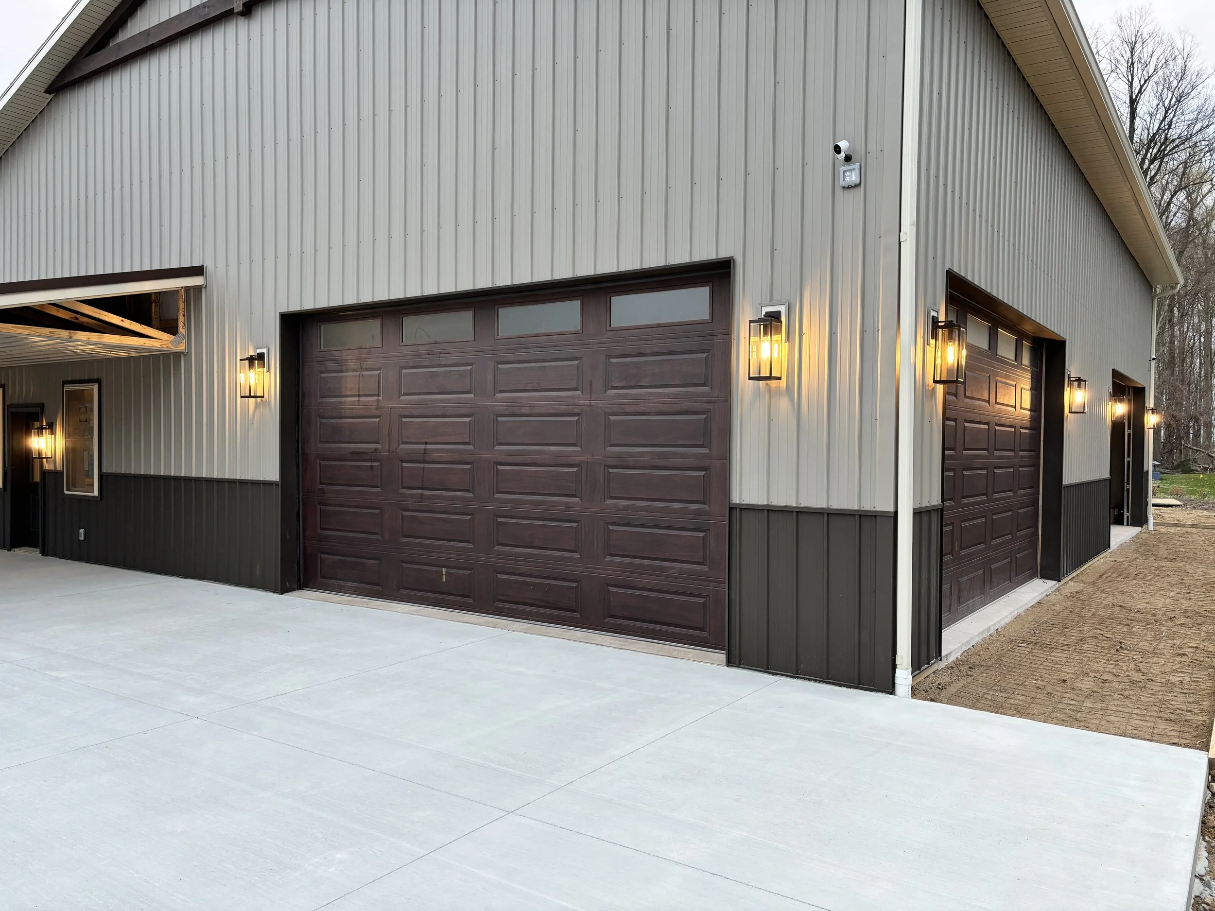 Exterior of a modern building with two large brown garage doors, metal siding, wall-mounted lights, and a security camera, with a concrete driveway and some trees in the background.