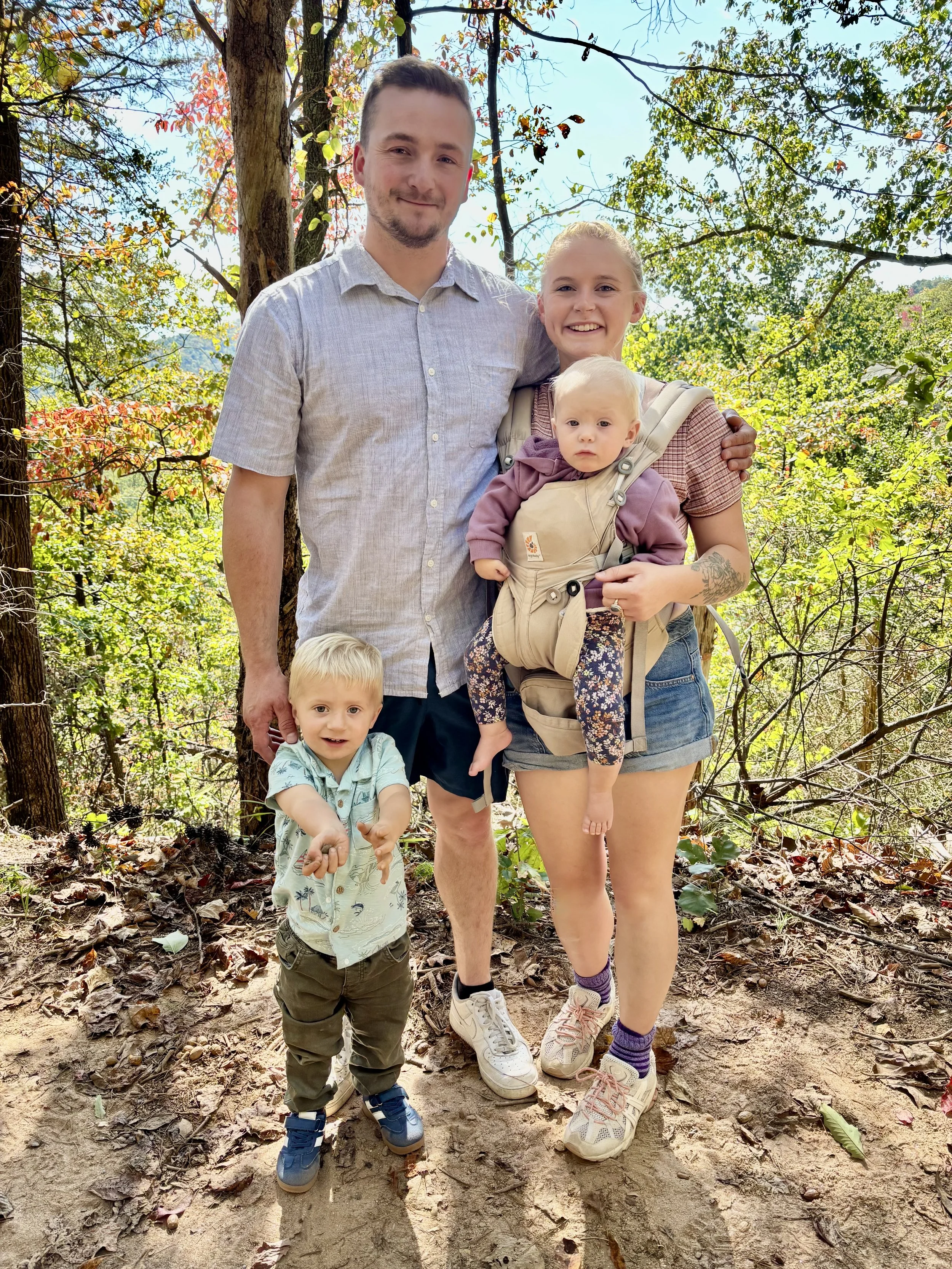 A family of four outdoors in a wooded area during autumn, smiling and posing for a photo. The family includes a man, a woman, a toddler boy, and a baby.