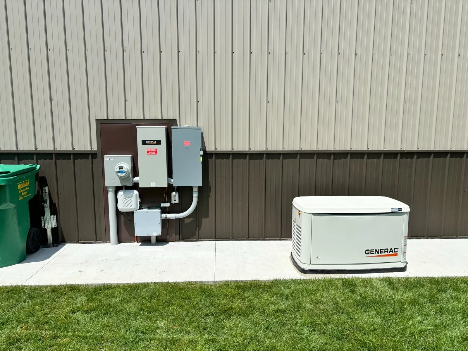 Exterior view of a beige and brown metal building wall with a generator and electrical box, a green trash bin, and a white portable generator on a concrete sidewalk with grass in the foreground.
