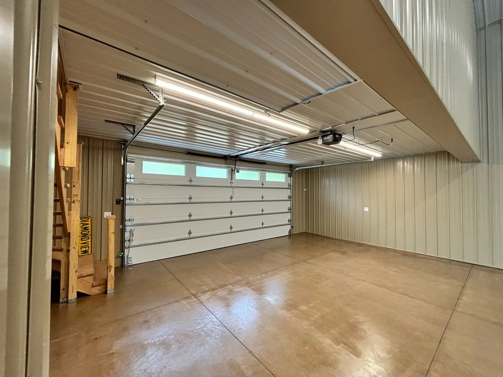 Empty garage with wooden flooring, beige paneled walls, white garage door, and ceiling with lighting and an automatic opener.