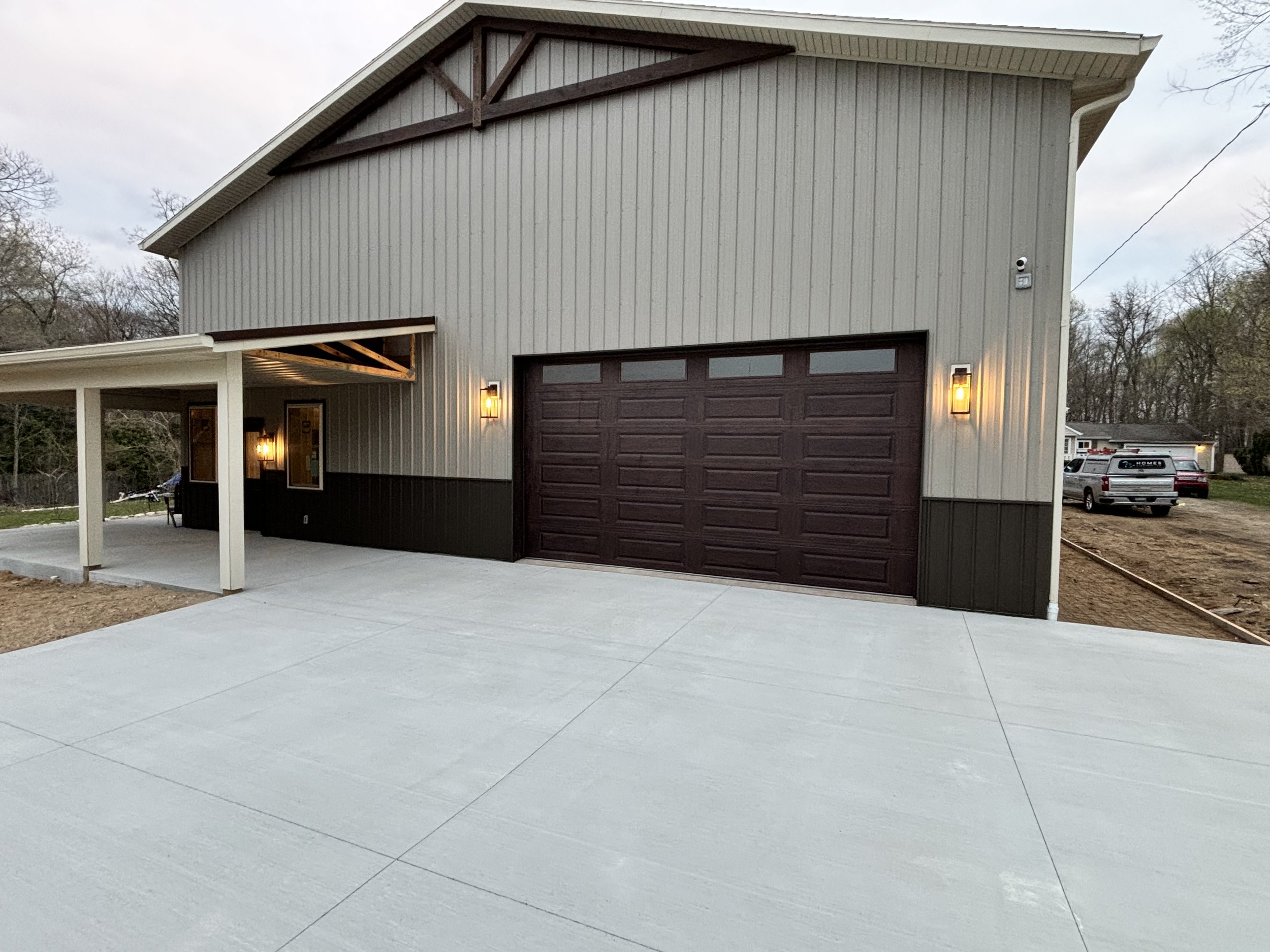 A newly completed two-story garage with abrown garage door, beige siding, two wall-mounted exterior lights, and a concrete driveway. The covered side porch has three windows and a ceiling with exposed wooden beams. The background shows a wooded area 