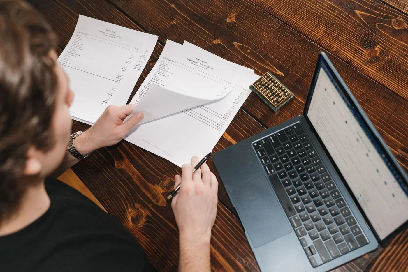 An over the shoulder photo from a male bookkeeper's perspective of his work station. He is flipping through ledger statements with a laptop and abacus nearby.