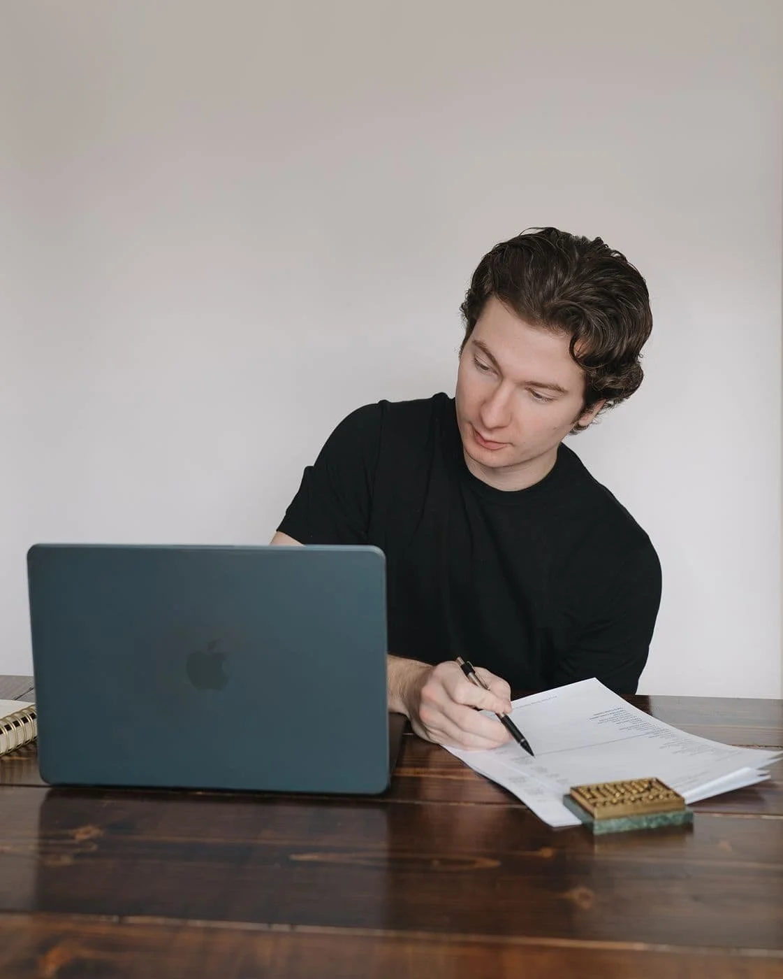 A male bookkeeper for small business working at a dark wood table, a laptop, papers and an abacus in front of him.