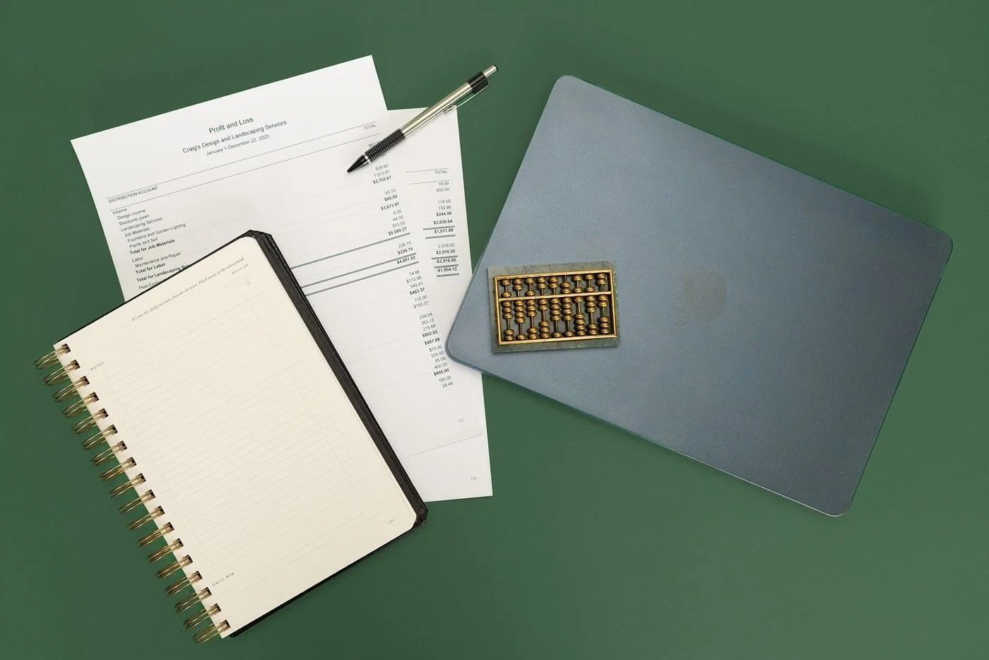 A top down detail photo of various bookkeeping items, including an abacus, a laptop, a notebook, and profit and loss statements, all sitting on a TrueBooks green background.