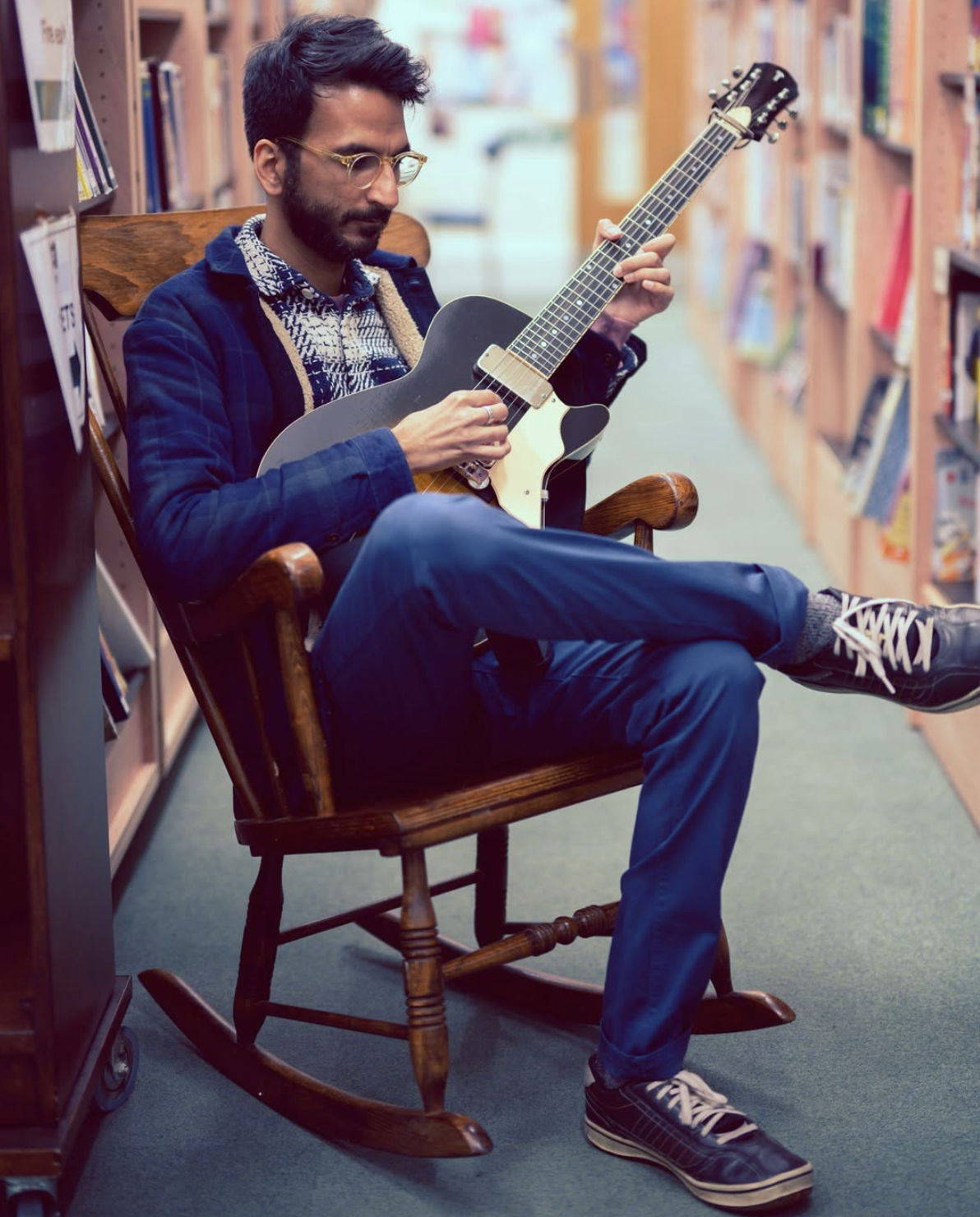 Amit Dattani playing an electric guitar while sitting in a wooden rocking chair in a bookstore.