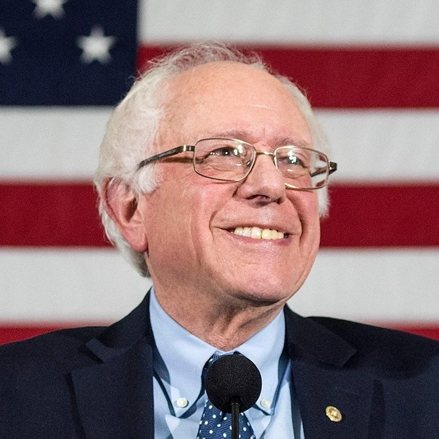 Senator Bernie Sanders smiling with glasses in a navy suit against an American flag background