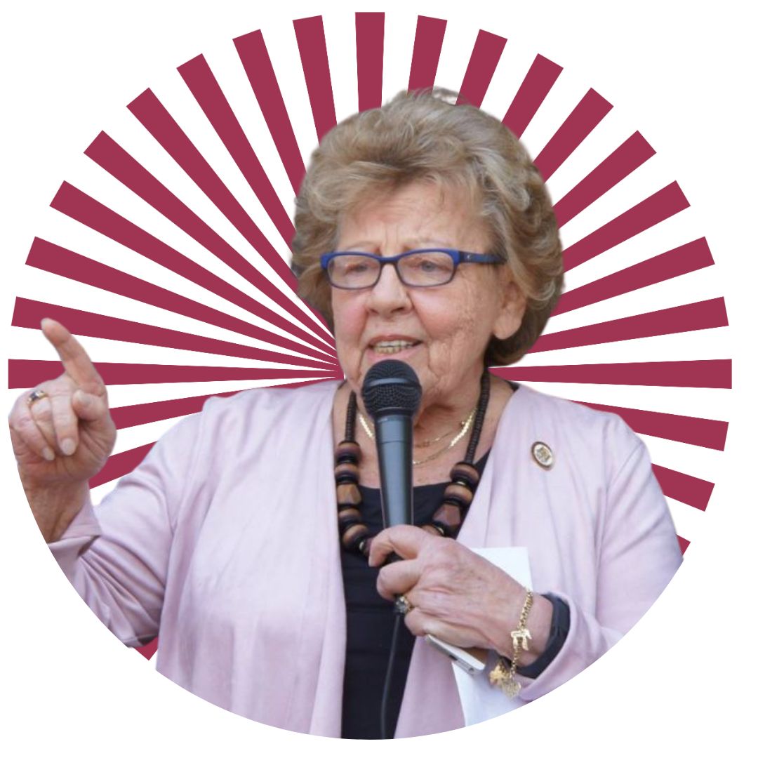 A Headshot of an elderly lady with short blond hair, glasses and a floral blazer, smiling against a blurred background.