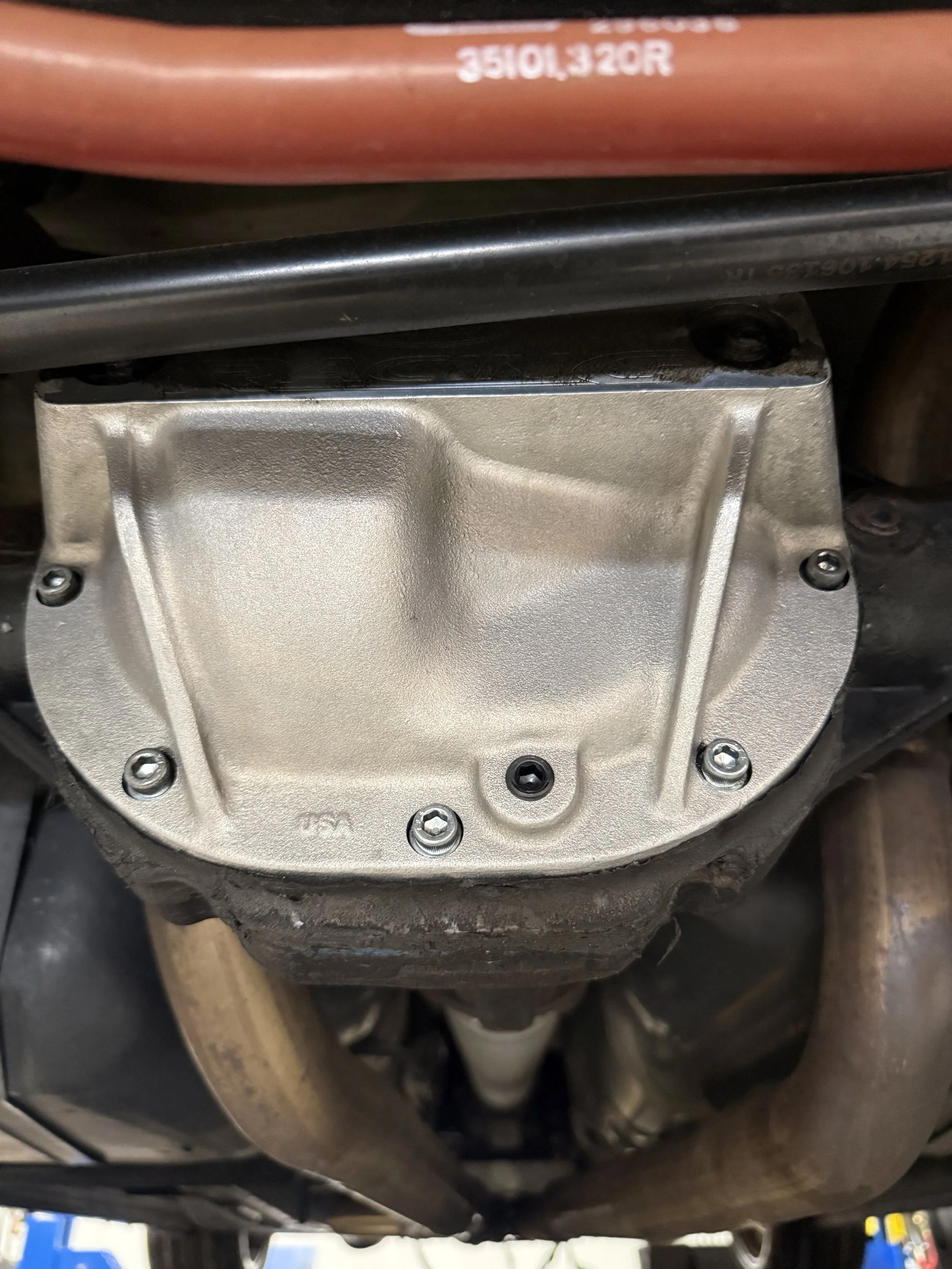 Close-up of a car engine component with a metallic cover secured by four screws, part of the vehicle's undercarriage with exhaust pipes visible underneath.