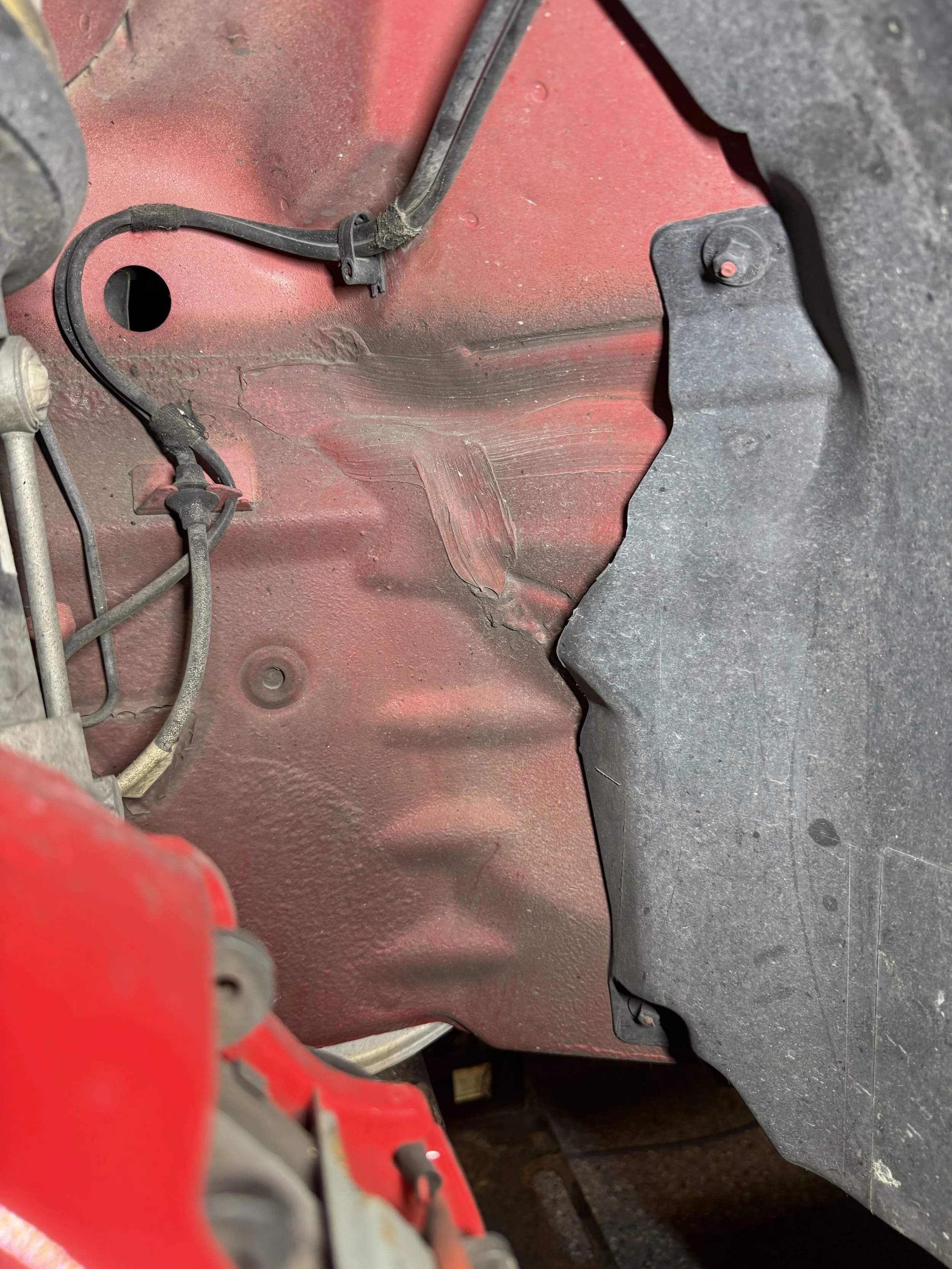 Close-up of the inside of a vehicle's wheel well with red and black painted surfaces, wires, and protective liners.