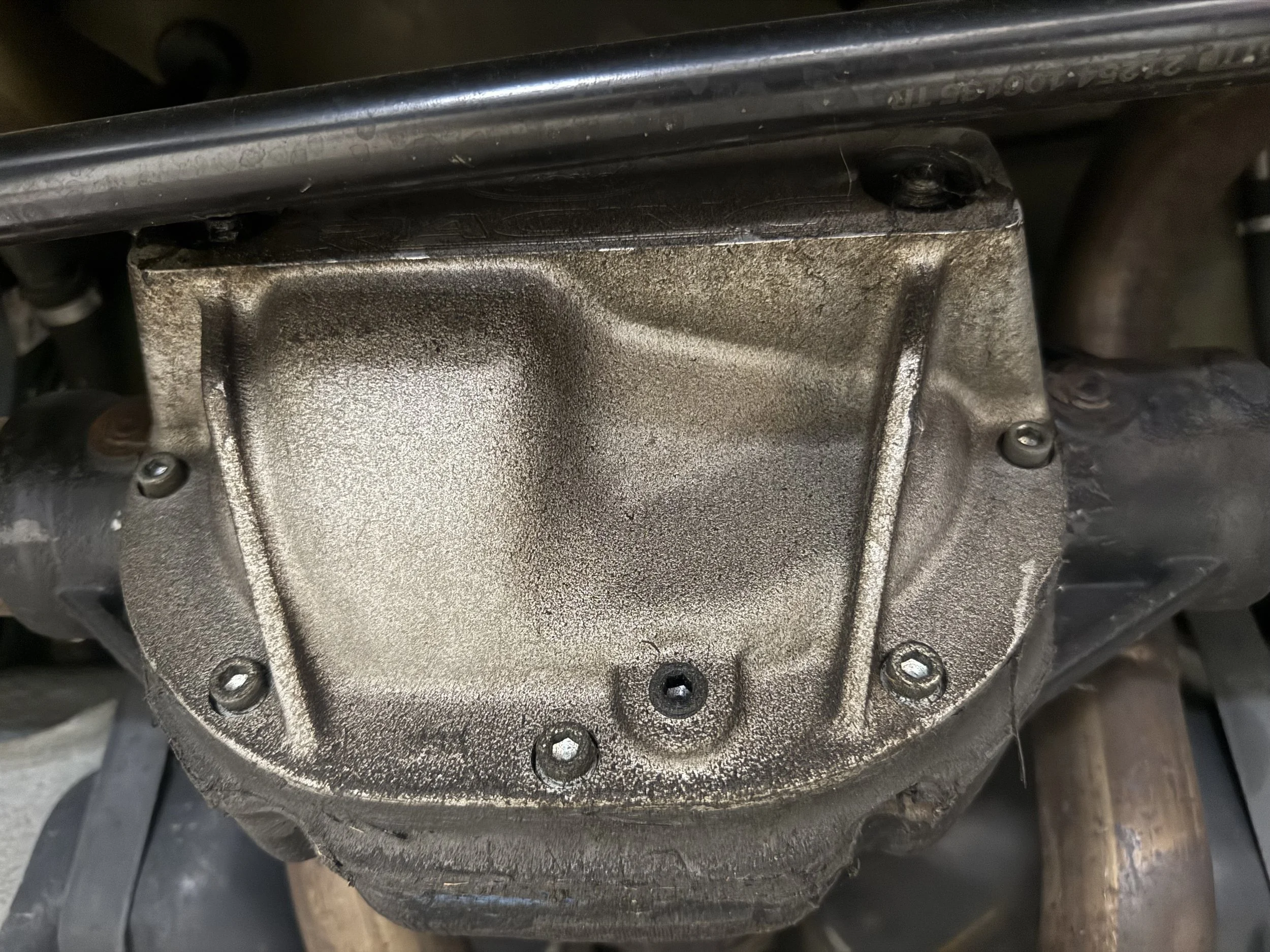 Close-up of a used engine or transmission component with a metallic, textured surface and six visible mounting bolts.