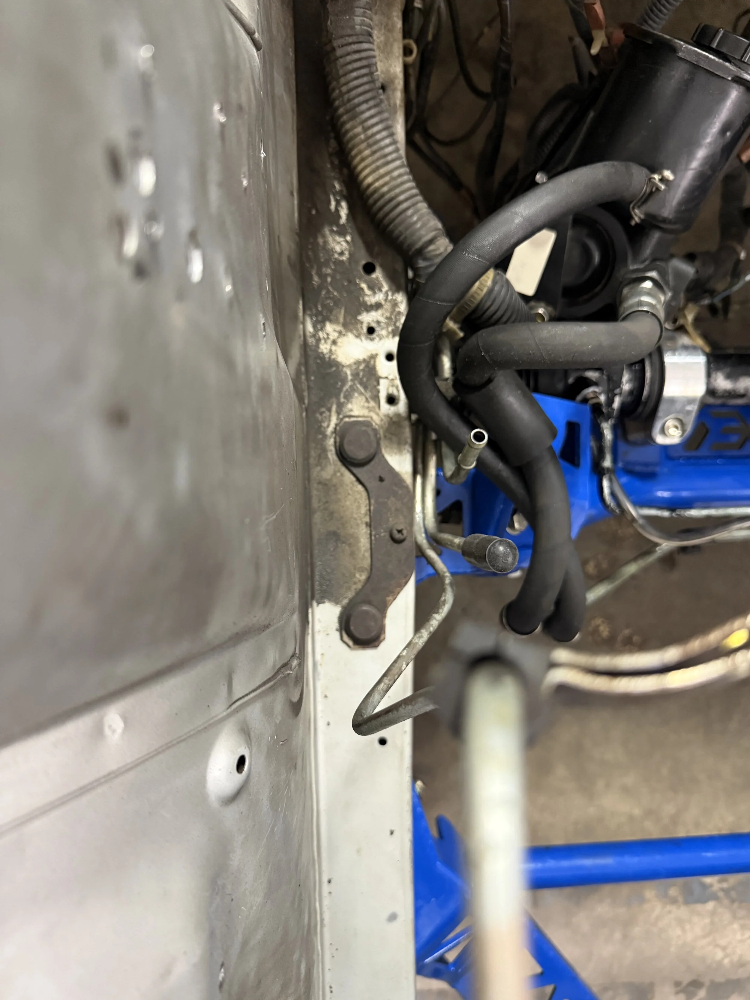 Close-up of an engine bay with hoses, wires, and metal components in focus, mounted to a metal surface, with some dirt and rust visible.