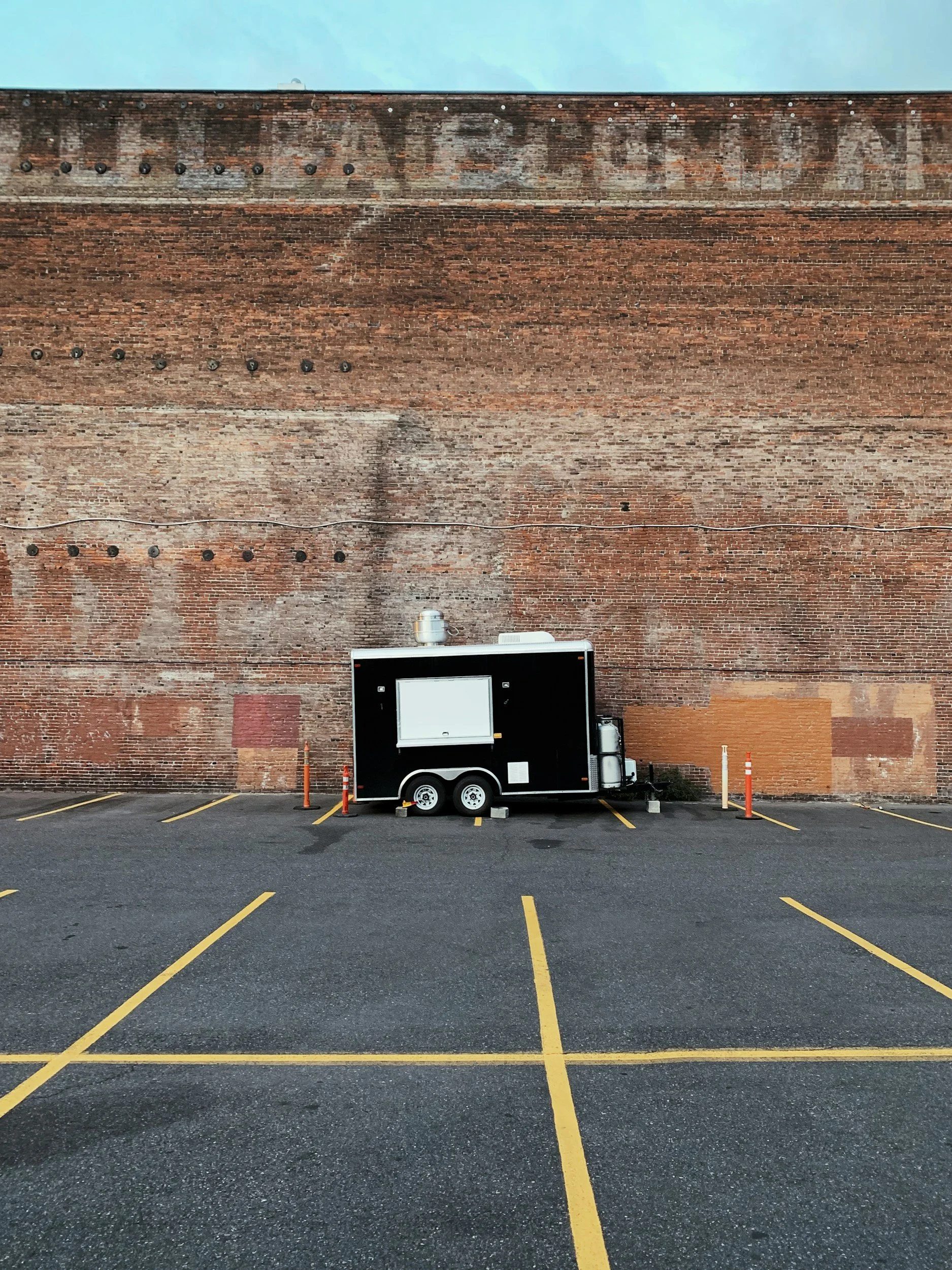 A black mobile food or concession trailer parked in an empty parking lot with yellow lines, against a tall brick wall.
