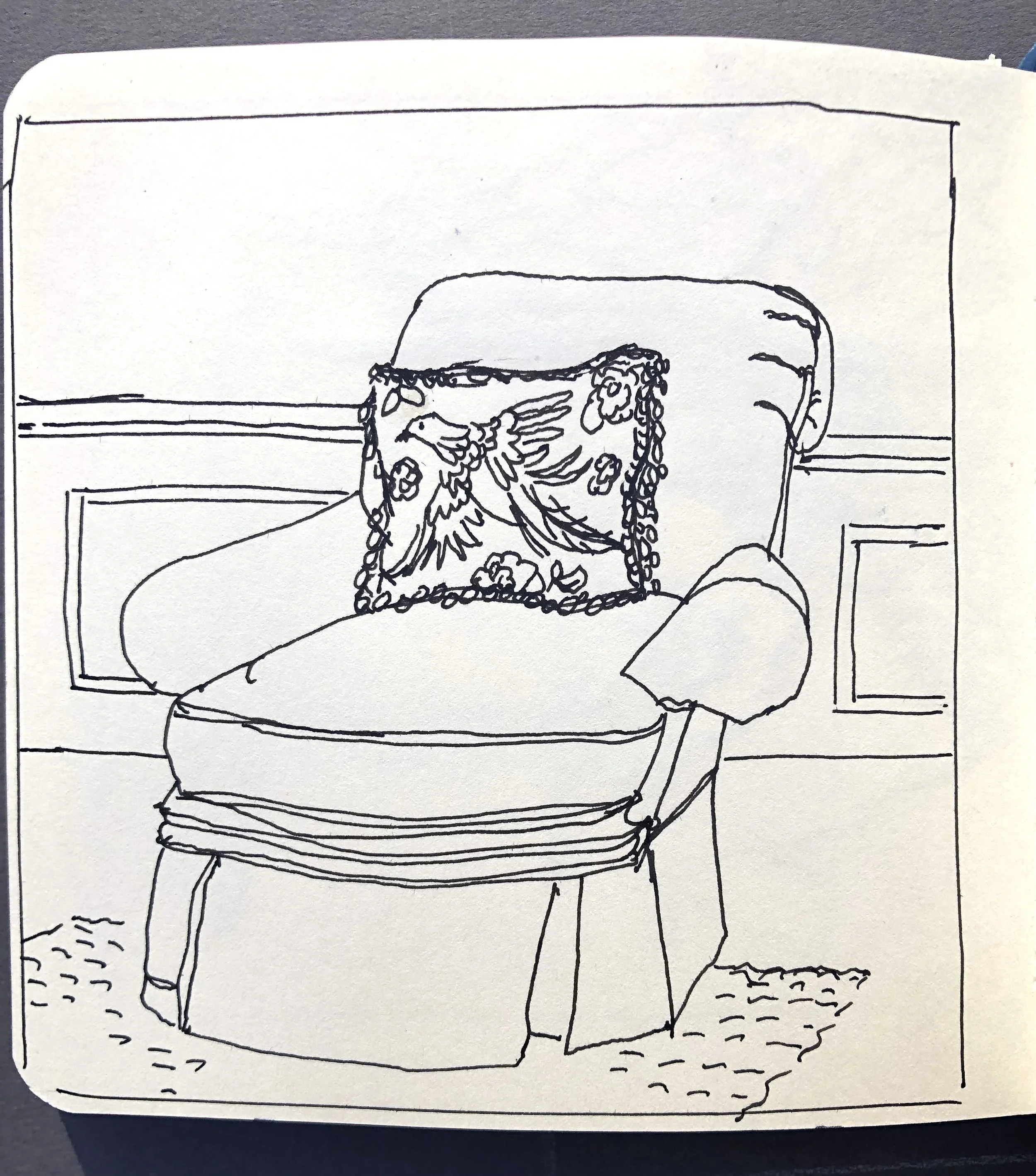 A line drawing of a chair with a decorative pillow featuring a bird and floral pattern, placed in front of a paneled wall.