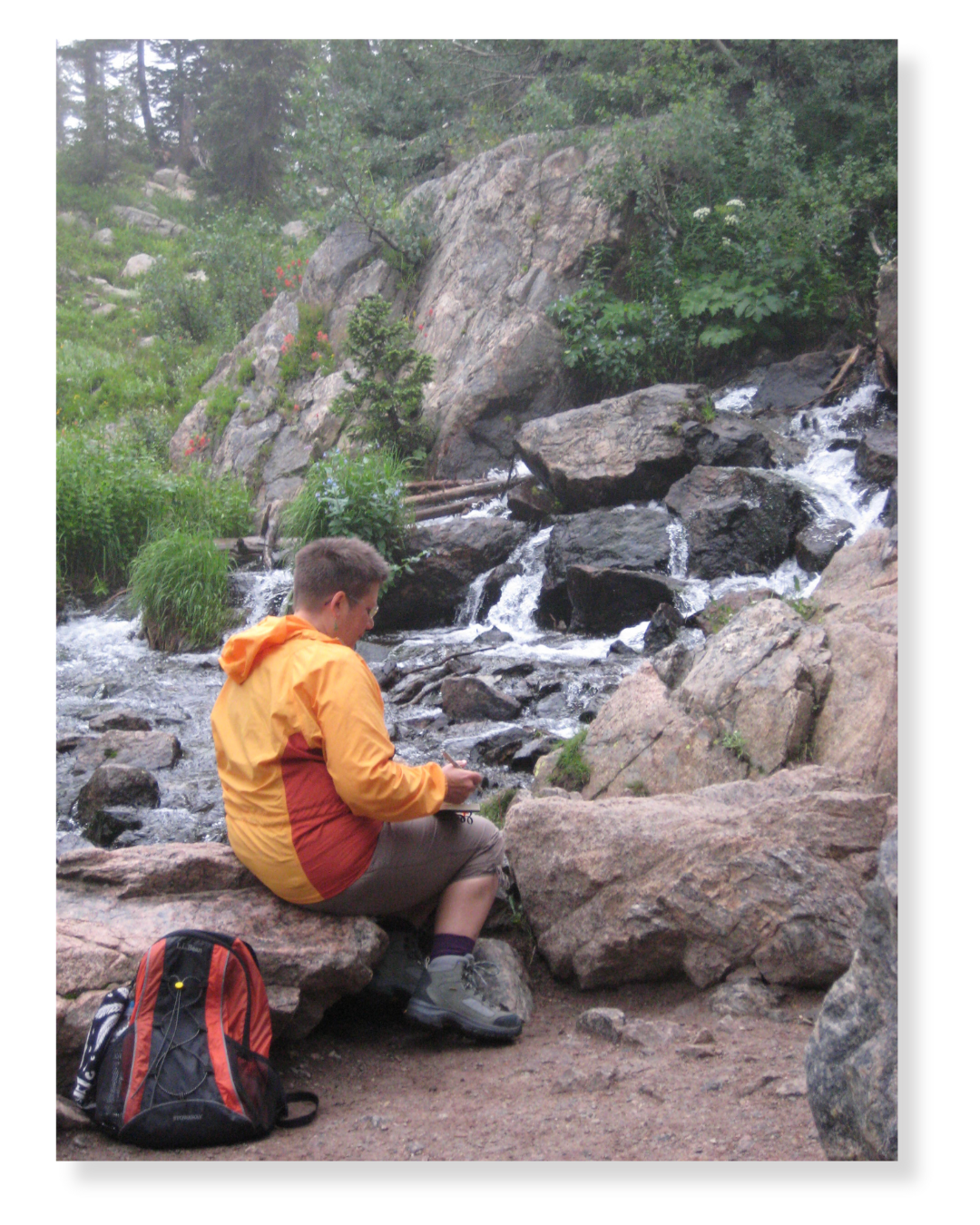 A person sitting on a large rock by a mountain stream, surrounded by greenery, while looking at something in their hands, with a backpack on the ground nearby.