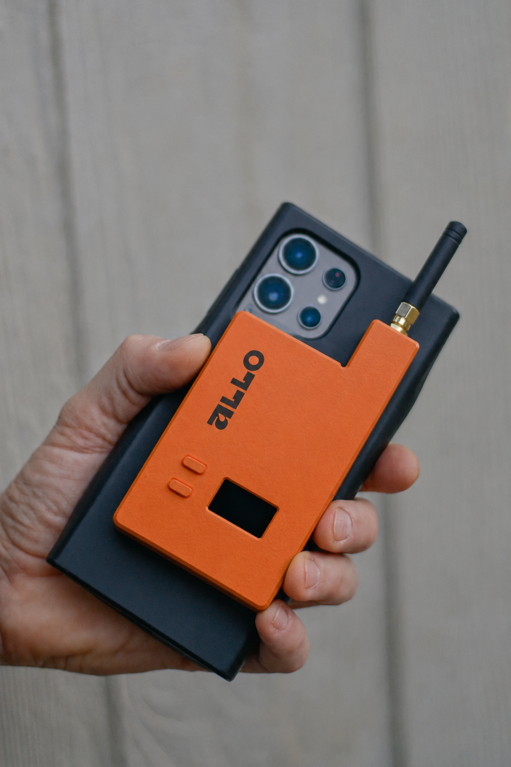 Persoon houdt een smartphone met een apparaat dat een GPS-antene. Op de telefoon rust een oranje apparaat met het woord 'OTLAP' erop, en een antenne. De achtergrond bestaat uit een lichte houten muur.