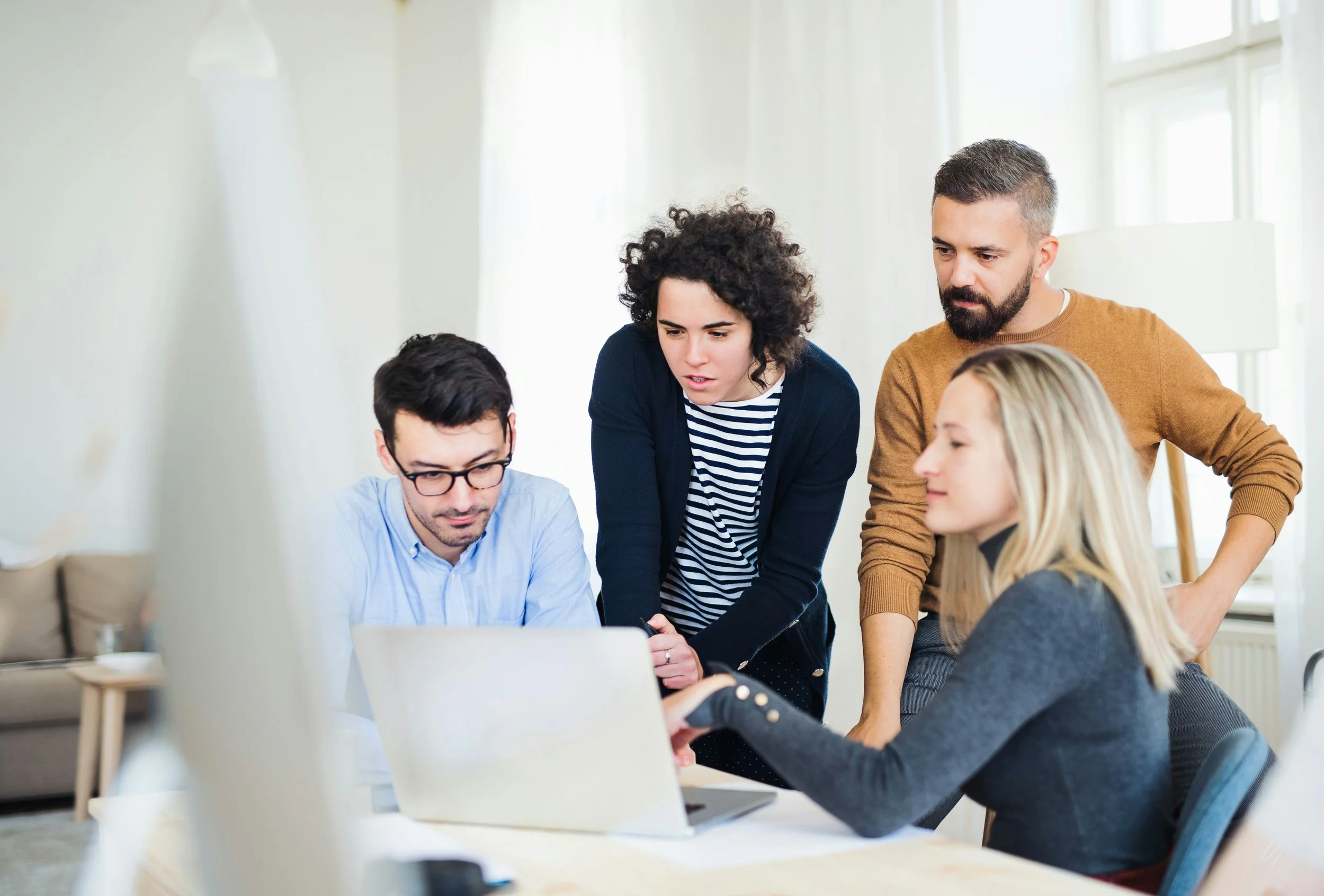 Group of four young adults collaborating around a laptop in a brightly lit room.
