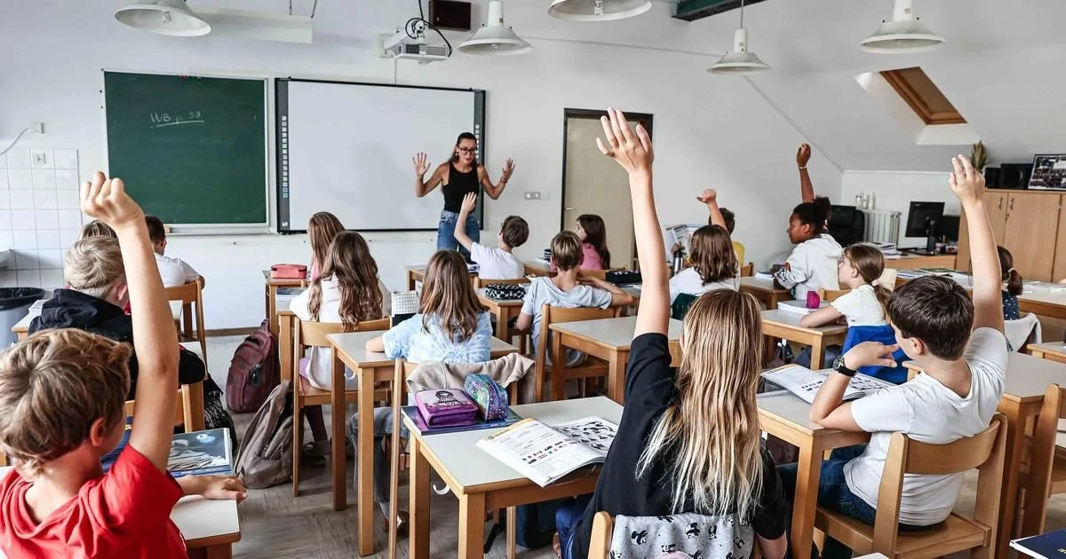 A classroom with students sitting at desks, raising hands to answer a question, and a teacher standing near a blackboard at the front.