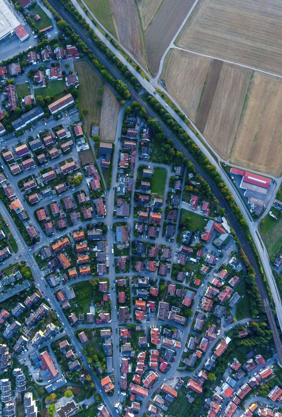 Luchtfoto van een woonwijk met huizen, straten en groene ruimtes, grenzend aan landbouwgrond en een spoorlijn.