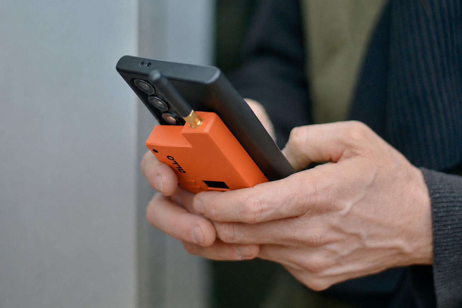 Een persoon vasthoudt een smartphone met een oranje apparaat met een antenne, mogelijk een detectie- of meetapparaat, verbonden met de smartphone.