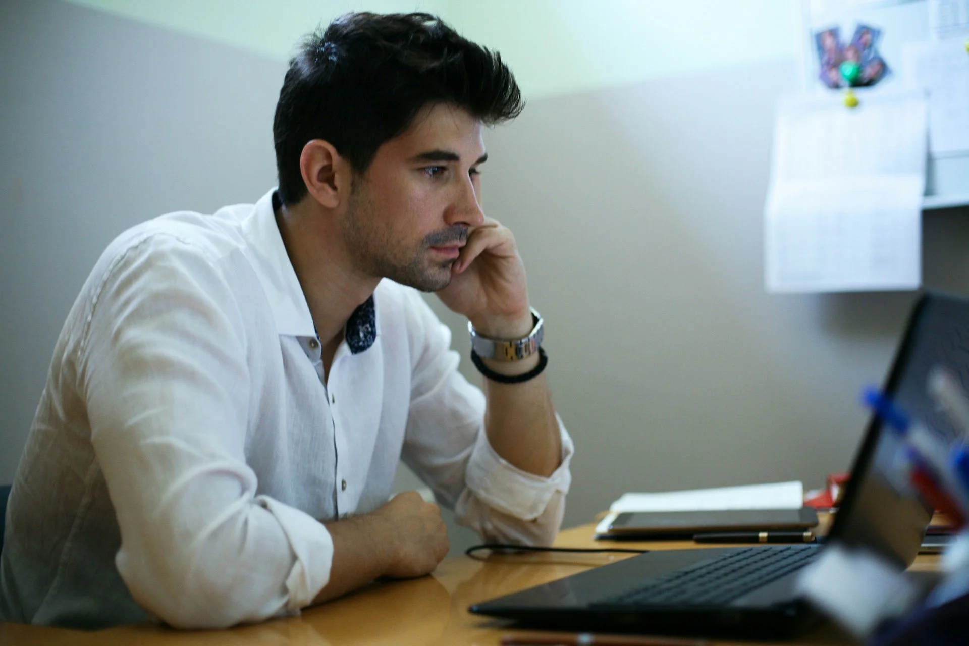 Een jonge man zit ernstig aan een bureau, zijn gezicht op zijn hand rustend, met een laptop, notitieboekje en papieren voor zich.