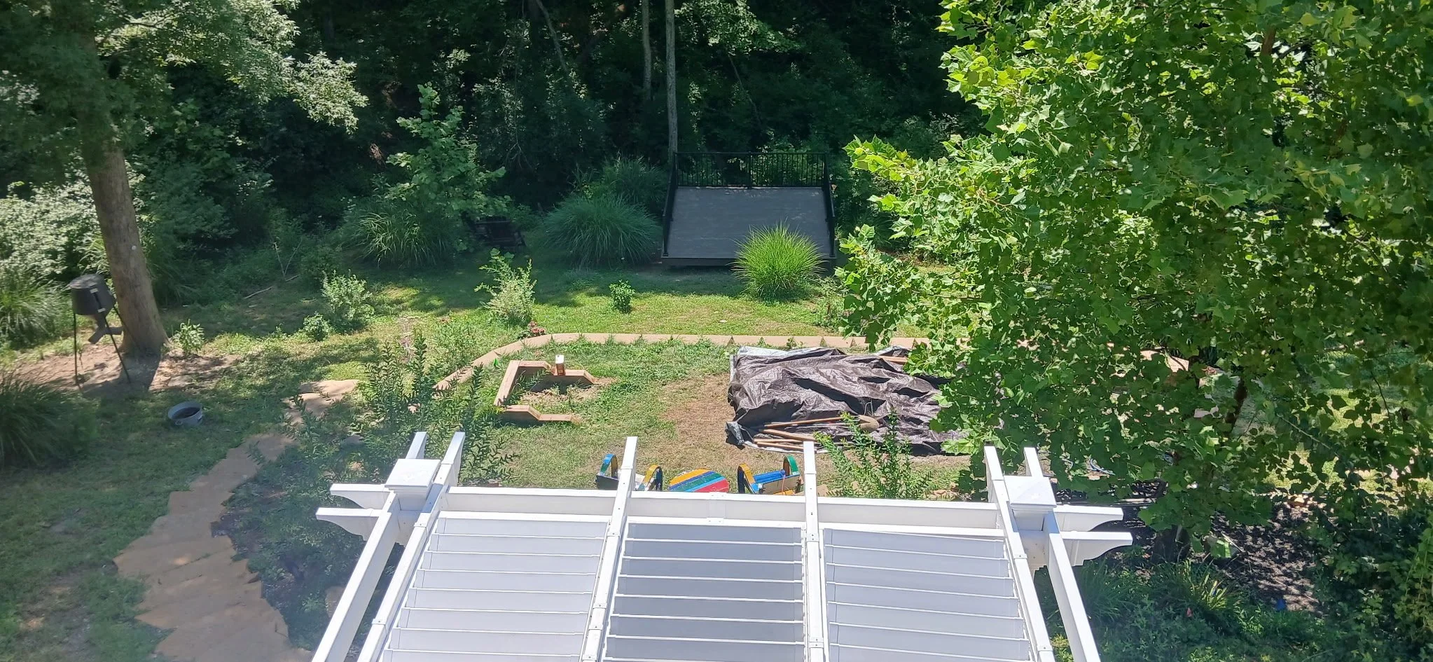 View from a second-story balcony looking down at a backyard with green grass, trees, and shrubs. There is a small firepit, a black tarp, a white deck railing, and a children's play set with colorful equipment. In the background, there is a black gymn