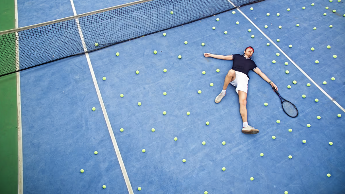 A man lying on a blue tennis court surrounded by numerous yellow tennis balls, holding a tennis racket, with a tennis net in the background.