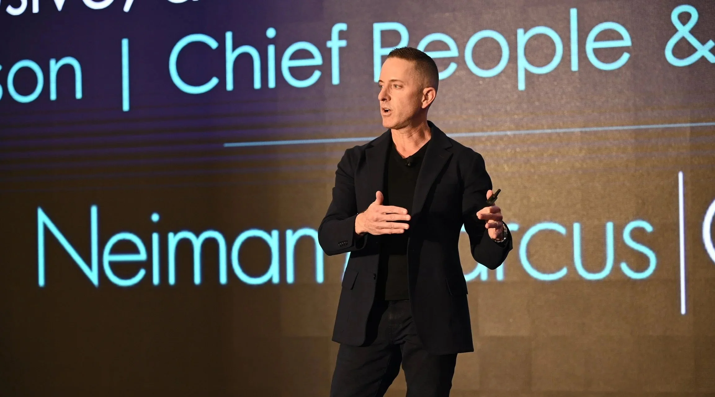 A man in a black suit giving a presentation on stage, with a large screen behind him displaying text.