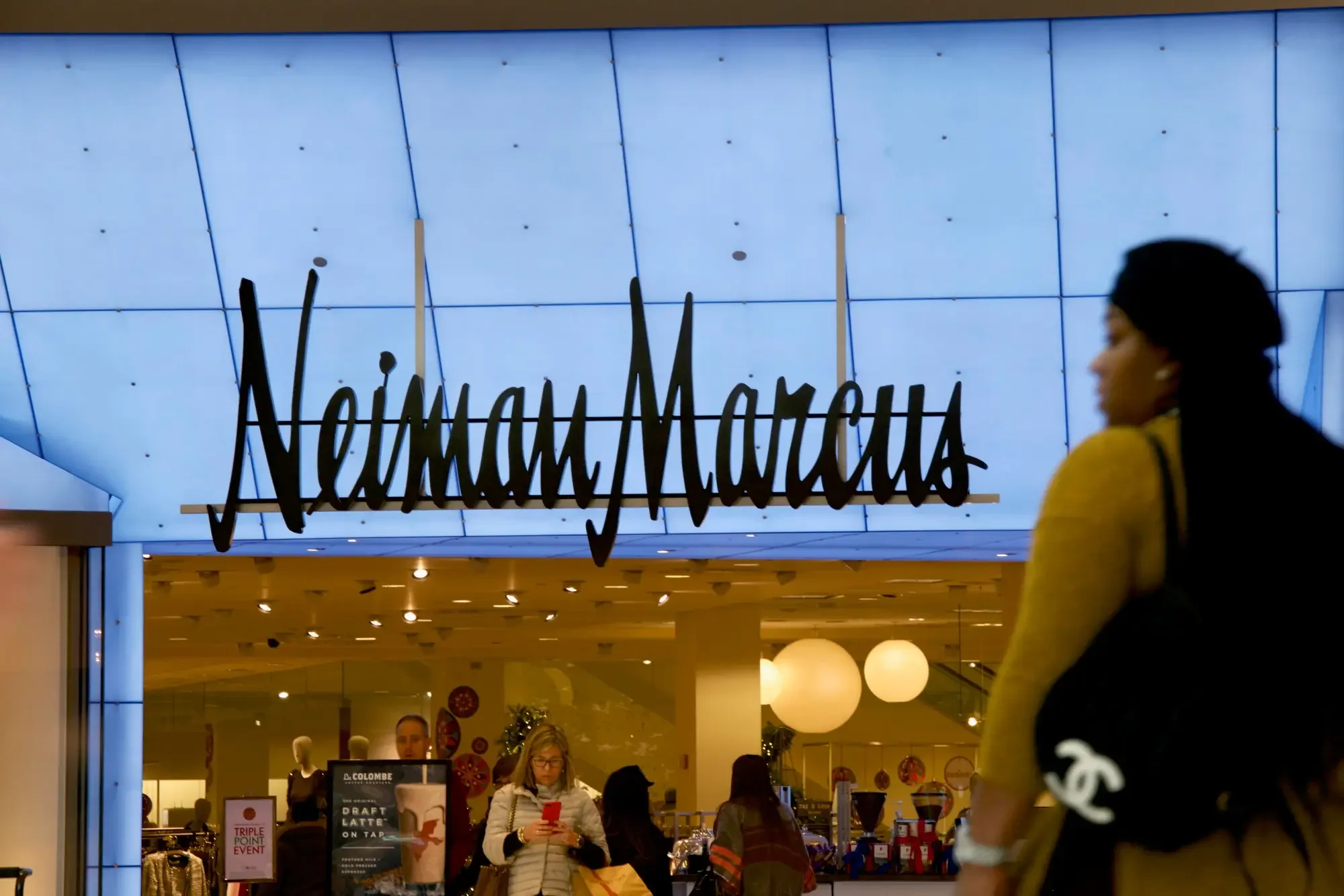 View of Neiman Marcus store entrance with a woman in yellow cardigan and black bag in foreground and shoppers inside, store sign visible overhead.